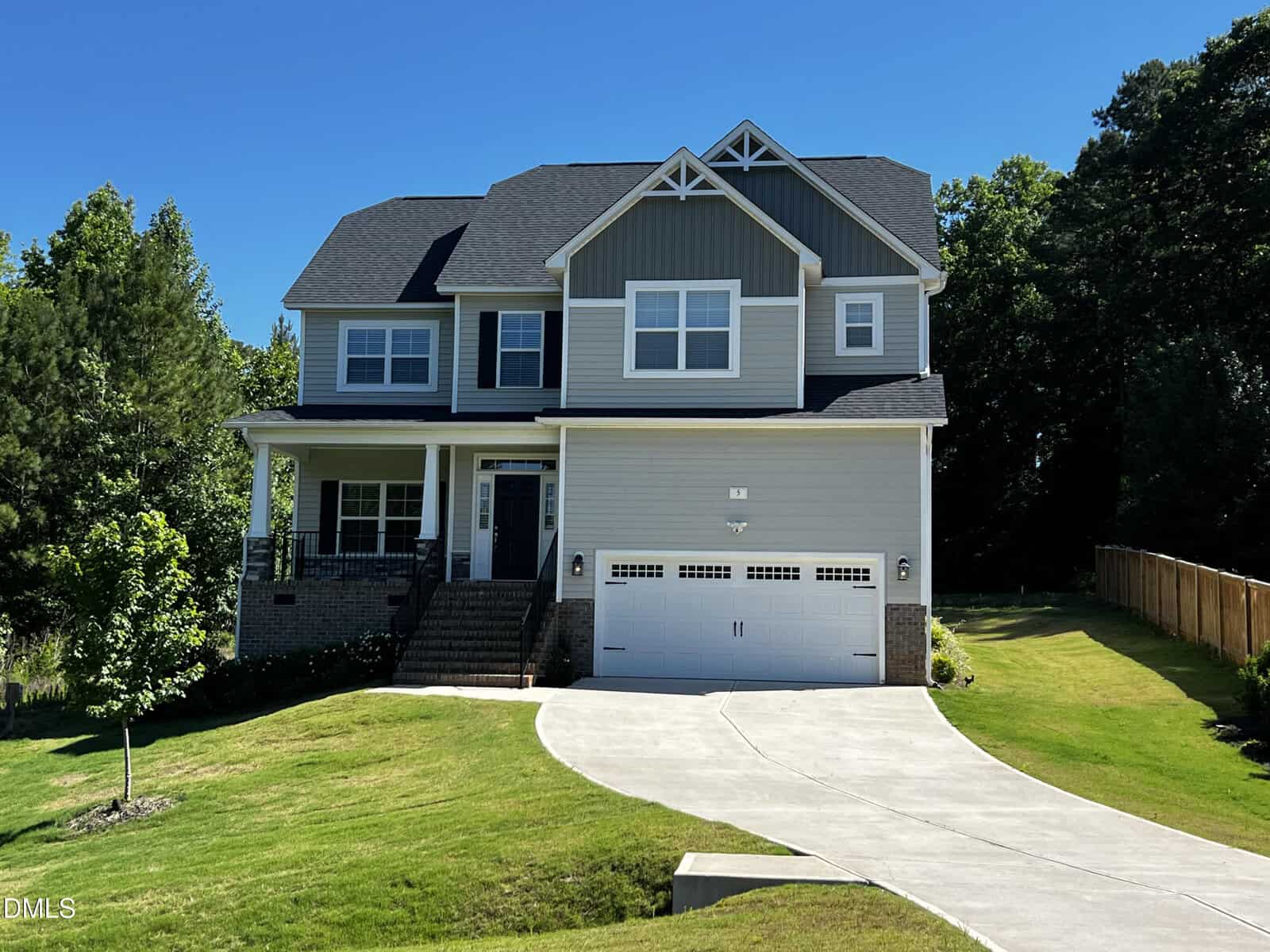 Two-story modern suburban house at 5 Cinnamon Teal Way, Youngsville, featuring gray siding, a covered porch, black roof, and a white two-car garage. Set on a sloped lawn with curved driveway, trees, and a wooden fence.