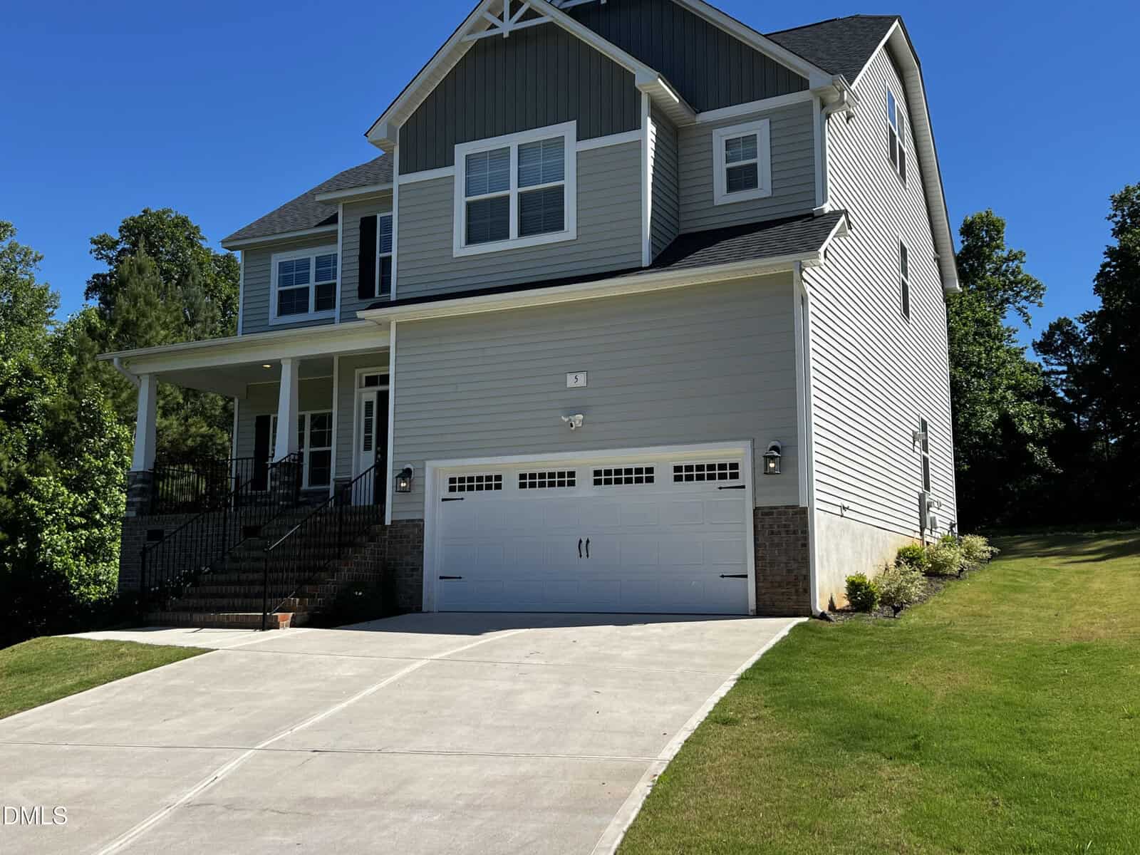 Two-story gray house with white trim, a front porch, and a double garage. The driveway is wide and clean, and the house is surrounded by green grass and trees under a clear blue sky.