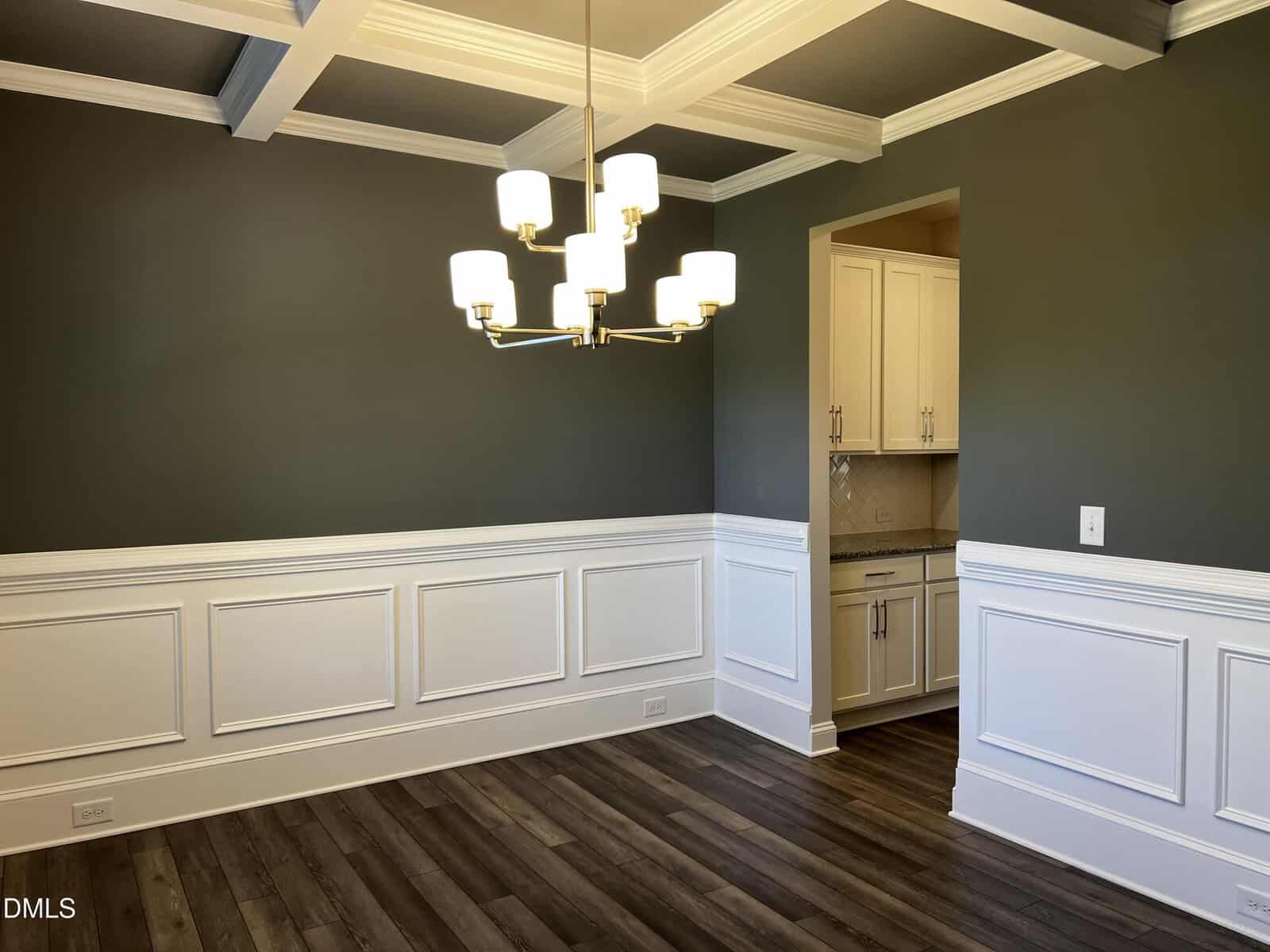 A dining room with dark green walls, white wainscoting, coffered ceiling, wood flooring, and a modern gold chandelier. An open doorway leads to a kitchen with white cabinets and granite countertops.