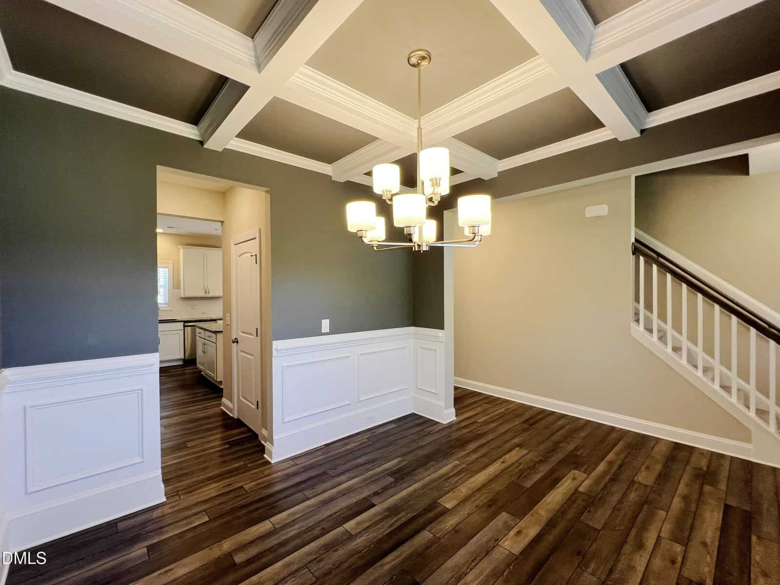 A modern dining room with dark wood floors, gray and white walls with wainscoting, a coffered ceiling, and a contemporary chandelier. An open doorway leads to a white kitchen, and a staircase is visible on the right.