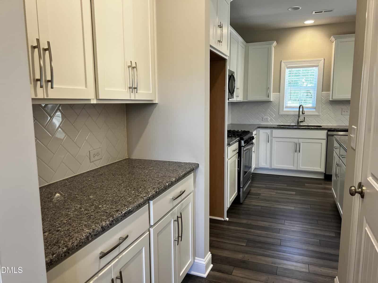 A modern kitchen with white cabinets, granite countertops, a herringbone tile backsplash, stainless steel appliances, dark wood floors, and a window above the sink letting in natural light.