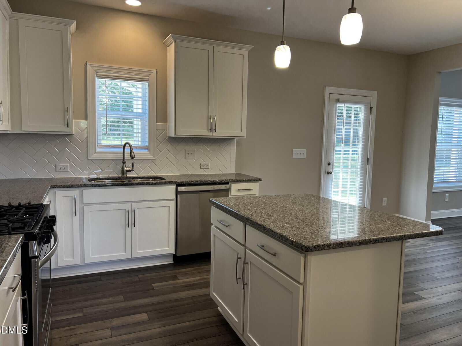 Modern kitchen with white cabinets, gray countertops, a center island, stainless steel appliances, tile backsplash, pendant lights, and large windows providing natural light. Door leads to a bright adjacent room.