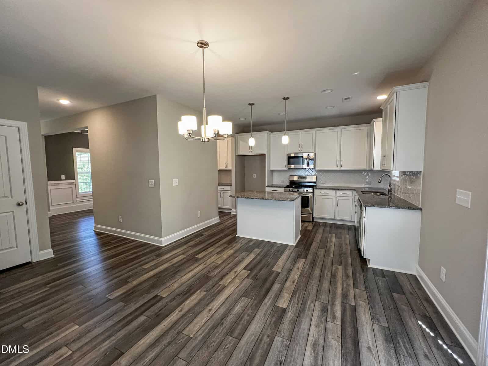 Modern kitchen and dining area with white cabinets, stainless steel appliances, pendant lights, wood-style flooring, and neutral-colored walls. Open layout with visible entry and natural light from a window in the adjacent room.