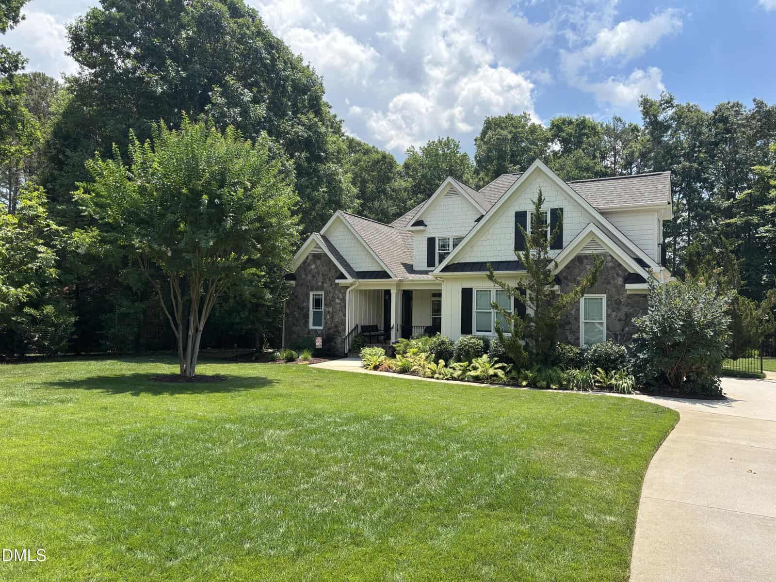 Two-story house with gray stone and white siding, black shutters, and a covered front porch, surrounded by green lawn, trees, shrubs, and a curved driveway on a sunny day.
