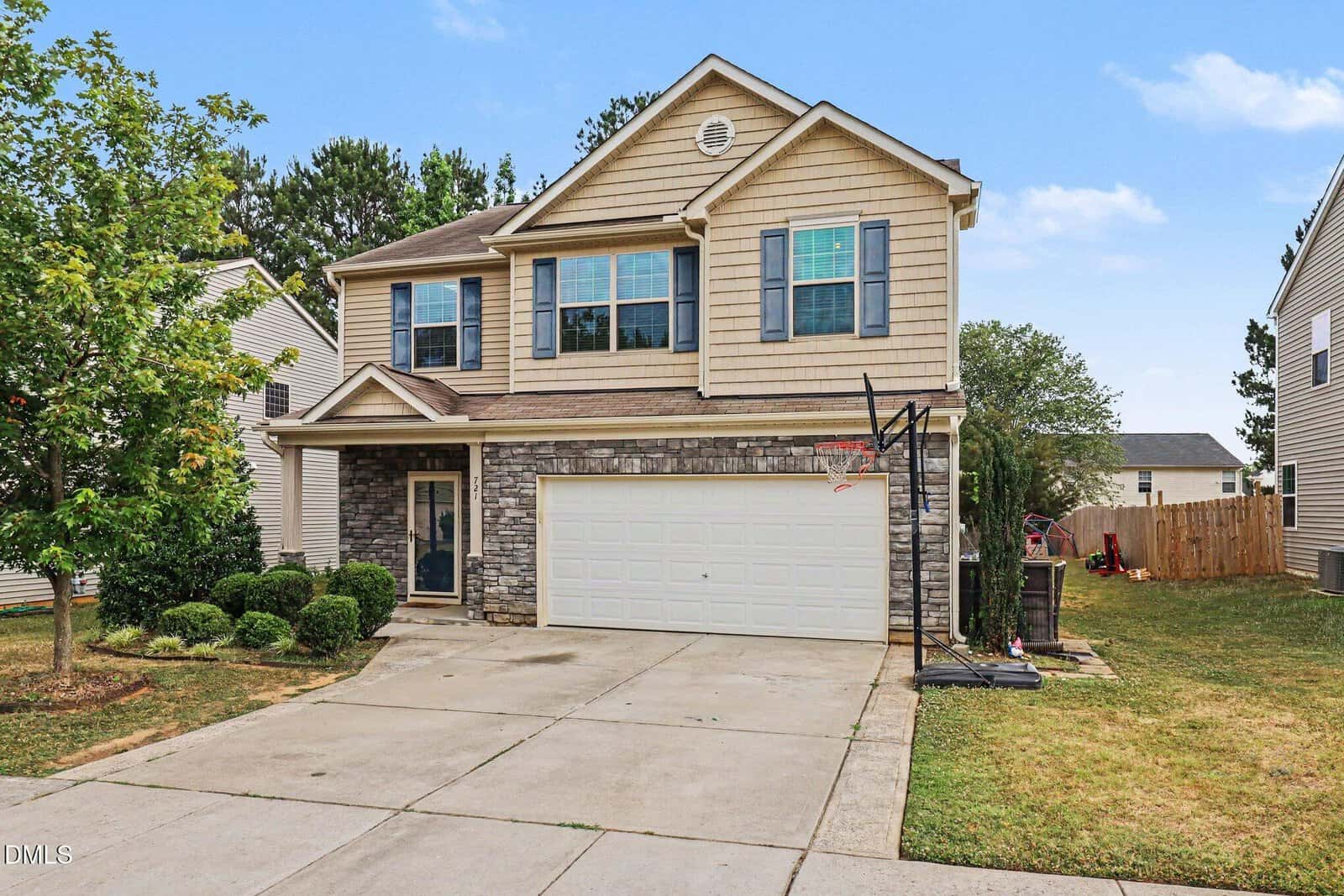 Charming two-story suburban house at 721 Tannerwell Avenue, Wake Forest, with beige siding, stone accent facade, double garage, and basketball hoop in the driveway; trimmed bushes and a small tree highlight the front yard amid neighboring homes.