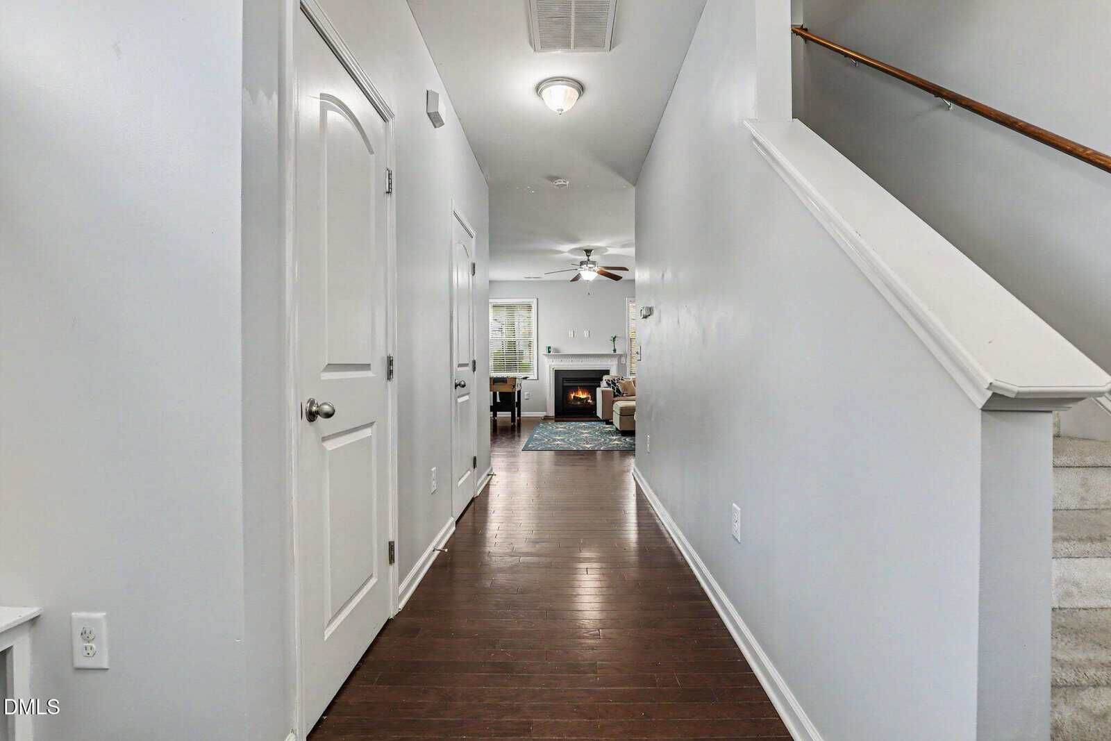 A hallway with hardwood floors, white walls, and a staircase to the right. At the end, there is a living room with a lit fireplace, ceiling fan, and a window with blinds.