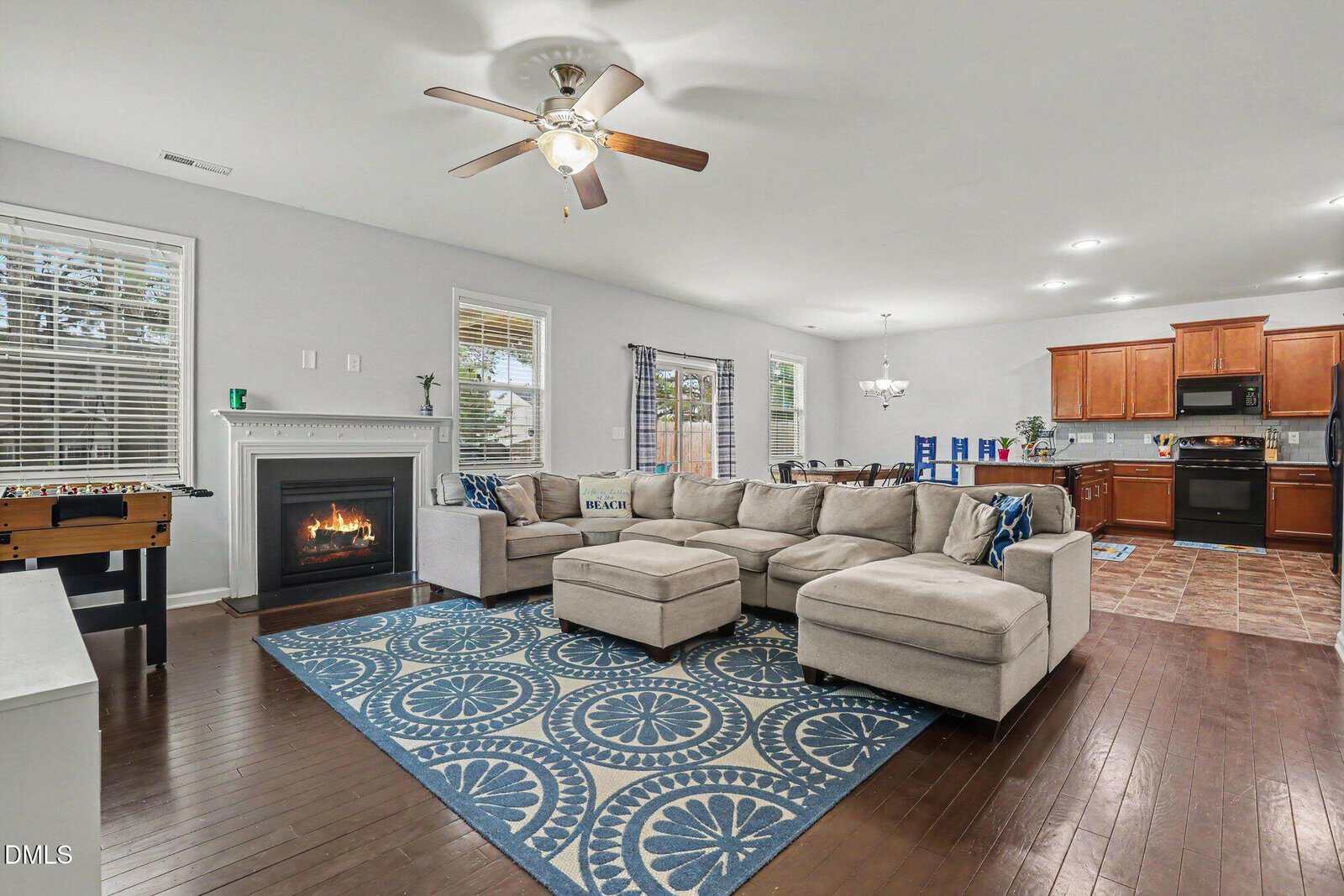 Spacious open-plan living room with a beige sectional sofa, blue patterned rug, fireplace, foosball table, and dining area. The kitchen with brown cabinets is in the background; large windows let in natural light.