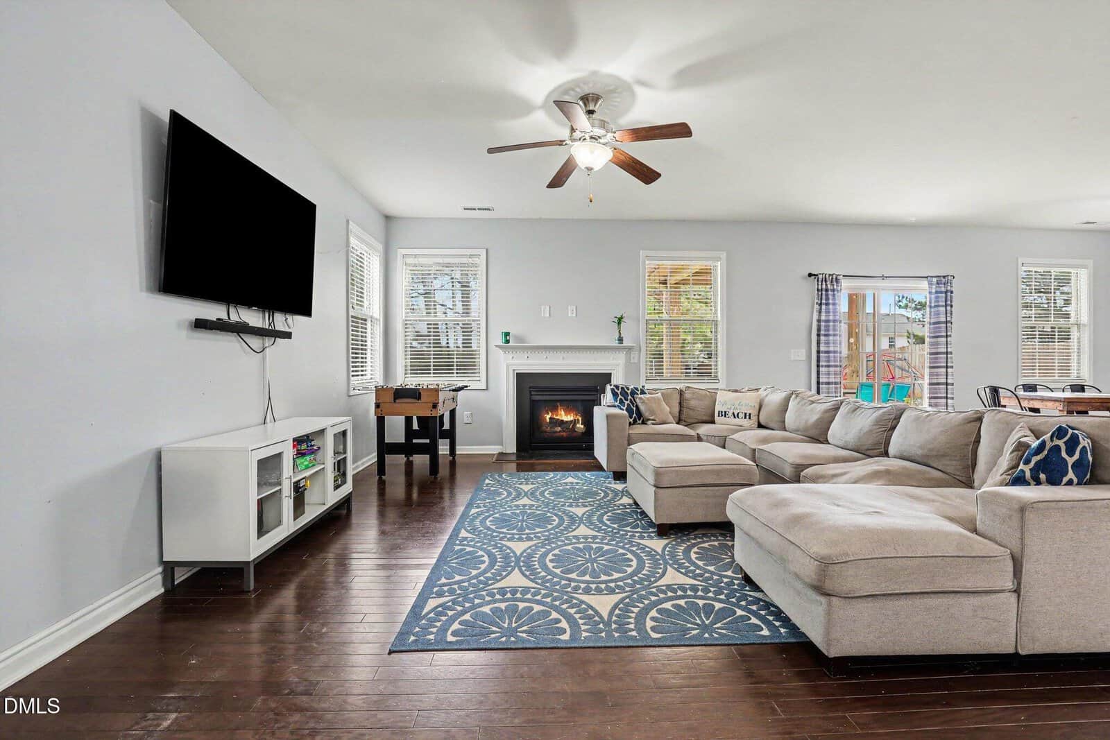 A spacious living room with a sectional sofa, patterned rug, wall-mounted TV, fireplace, and large windows letting in natural light. Hardwood floors and a dining area are visible in the background.