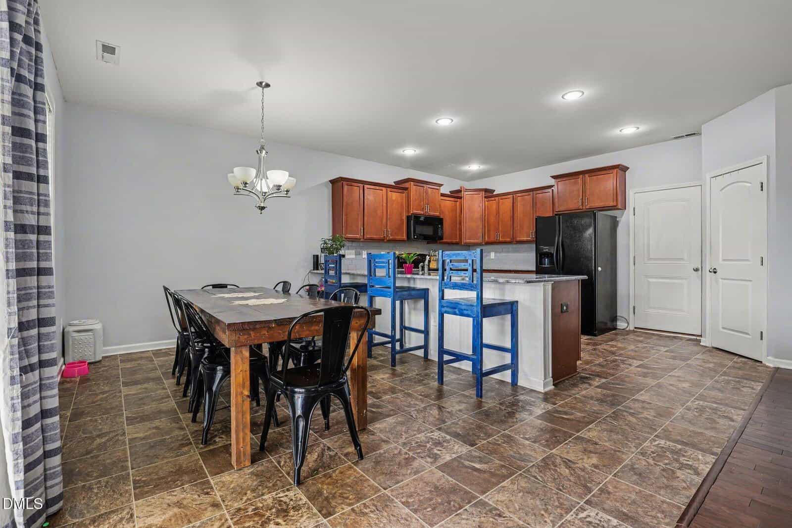 Spacious kitchen and dining area with brown tile flooring, wooden cabinets, black appliances, a large wooden dining table with black chairs, and a kitchen island with blue barstools. Light gray walls and modern lighting.
