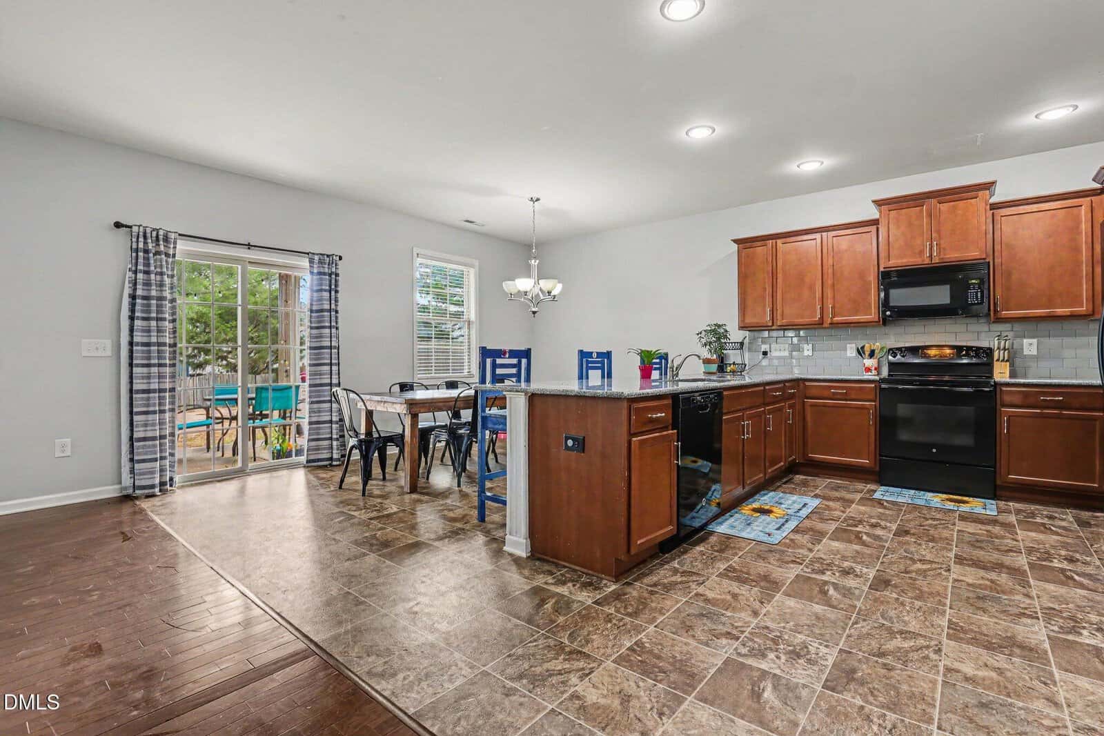 Open kitchen and dining area with wood cabinets, black appliances, island, tile floor, dining table with metal chairs, and sliding glass doors leading to a backyard with a playset visible outside.