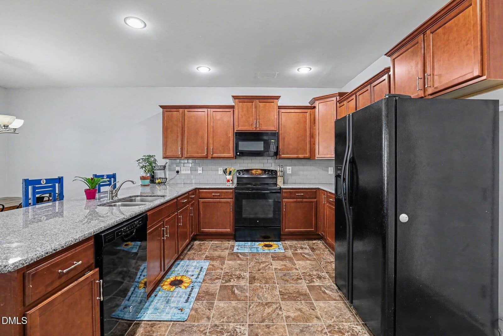 Modern kitchen with brown cabinets, black appliances, granite countertops, double sink, tile floor, and bright lighting. Two blue sunflower rugs, a plant, and colorful chairs add accents.