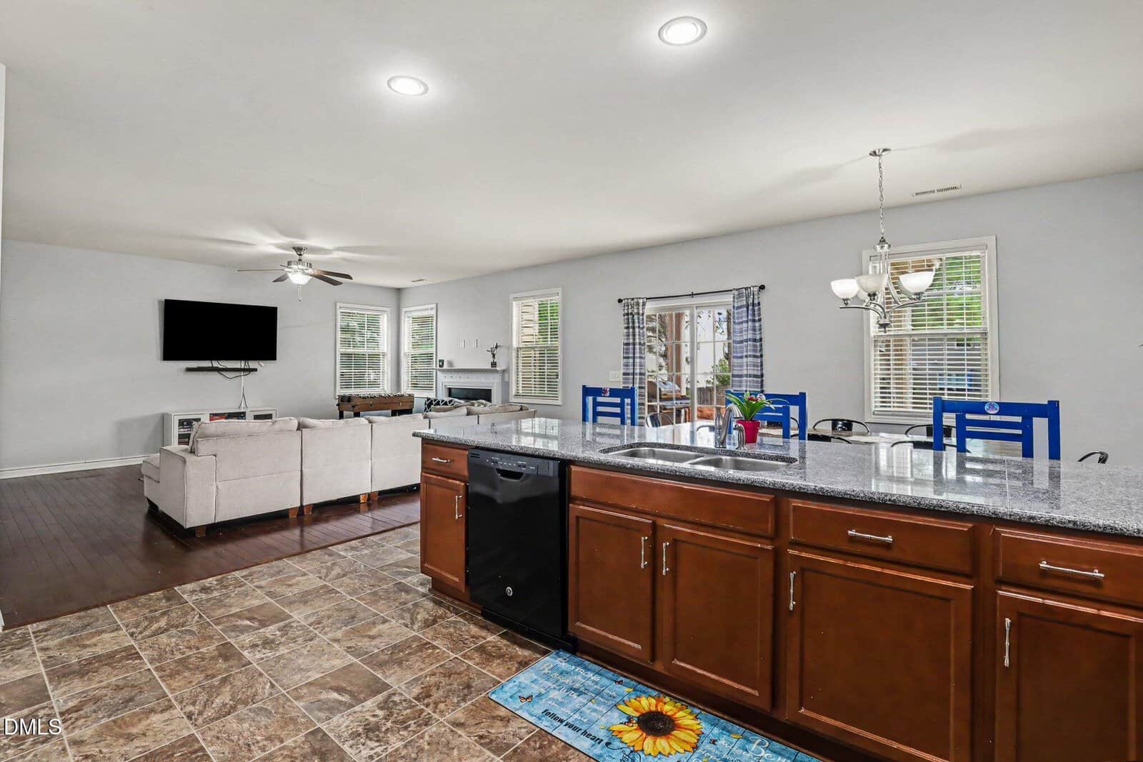 Spacious open-concept kitchen and living area with brown cabinets, granite countertops, tiled floor, and a living room with a sectional sofa, TV, and large windows letting in natural light.