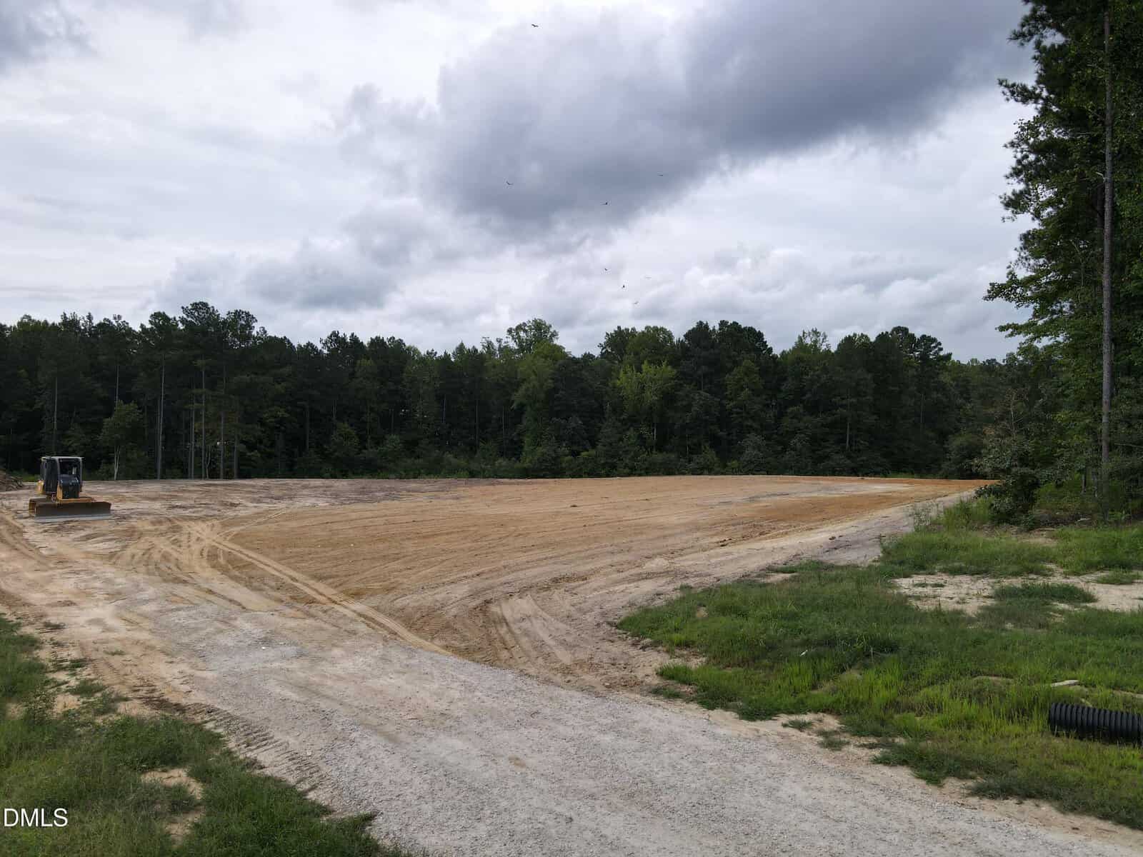 A cleared, sandy lot at 105 Mass Way in Zebulon is bordered by trees under a cloudy sky. A construction vehicle is parked on the left side, and tire tracks are visible on the ground. Dense forest surrounds the open area.