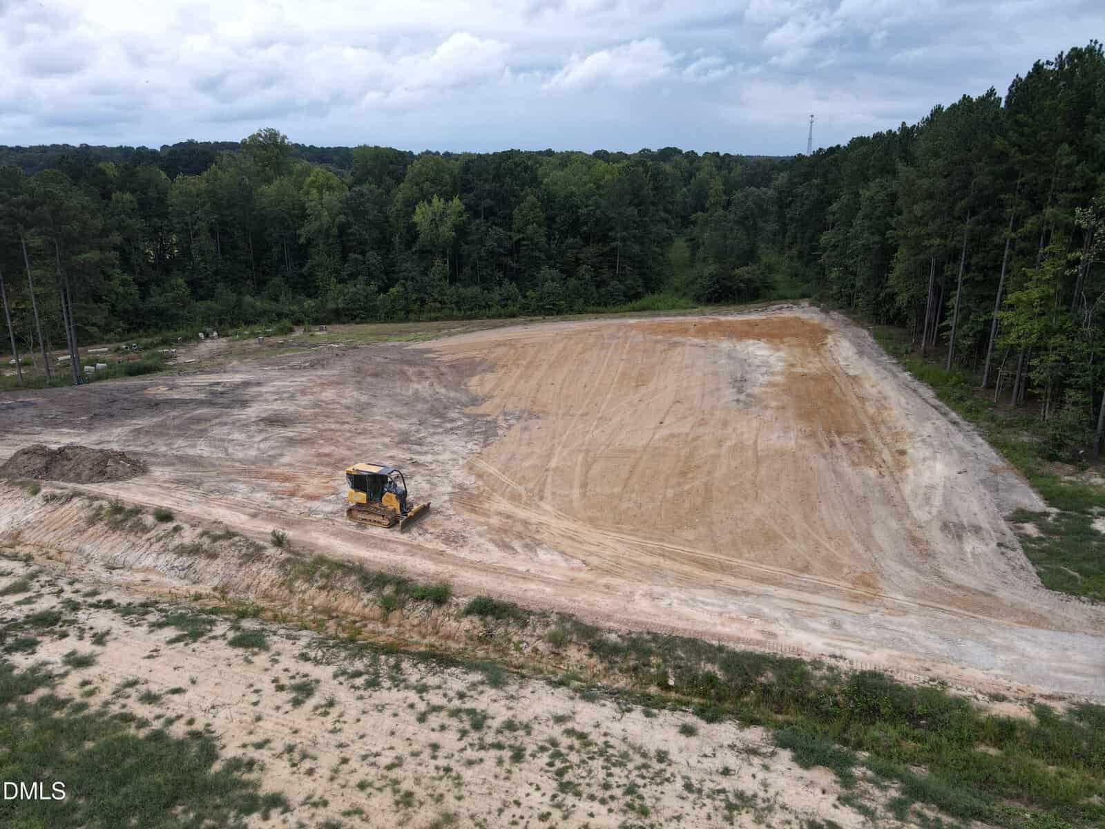 A bulldozer is parked on a large, cleared area of dirt surrounded by trees, with cloudy skies above.
