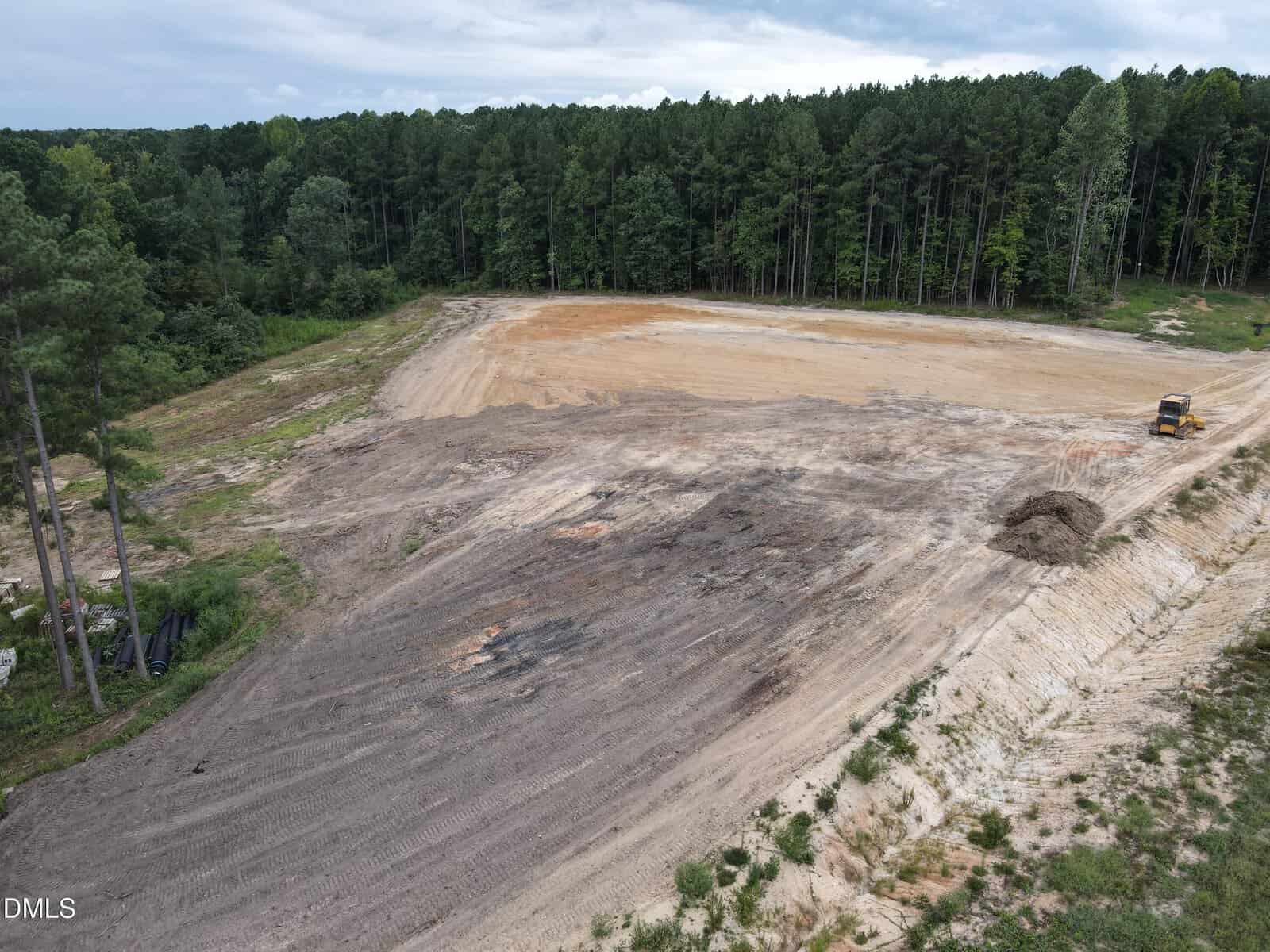 A construction site with cleared, leveled ground bordered by forest. A truck is parked near the cleared area. The sky is partly cloudy.