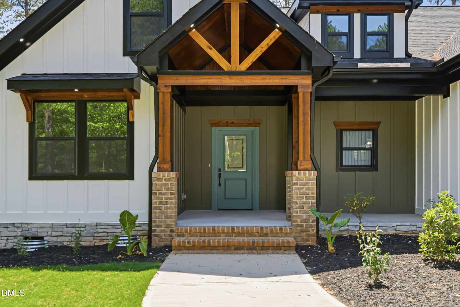 A modern house entryway with a green front door, brick steps, wooden beams, and white and green vertical siding. Small plants and shrubs line the landscaped garden in front.