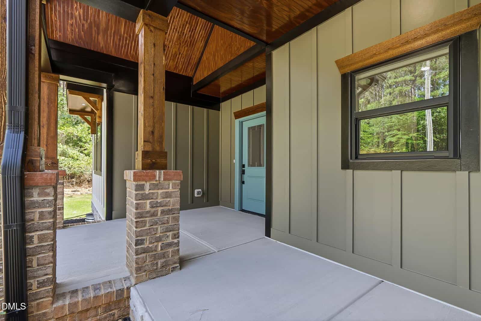 Covered modern porch with wooden columns, brick base, green siding, a blue front door, and a window showing trees outside. Concrete floor and ceiling feature wood paneling accents.