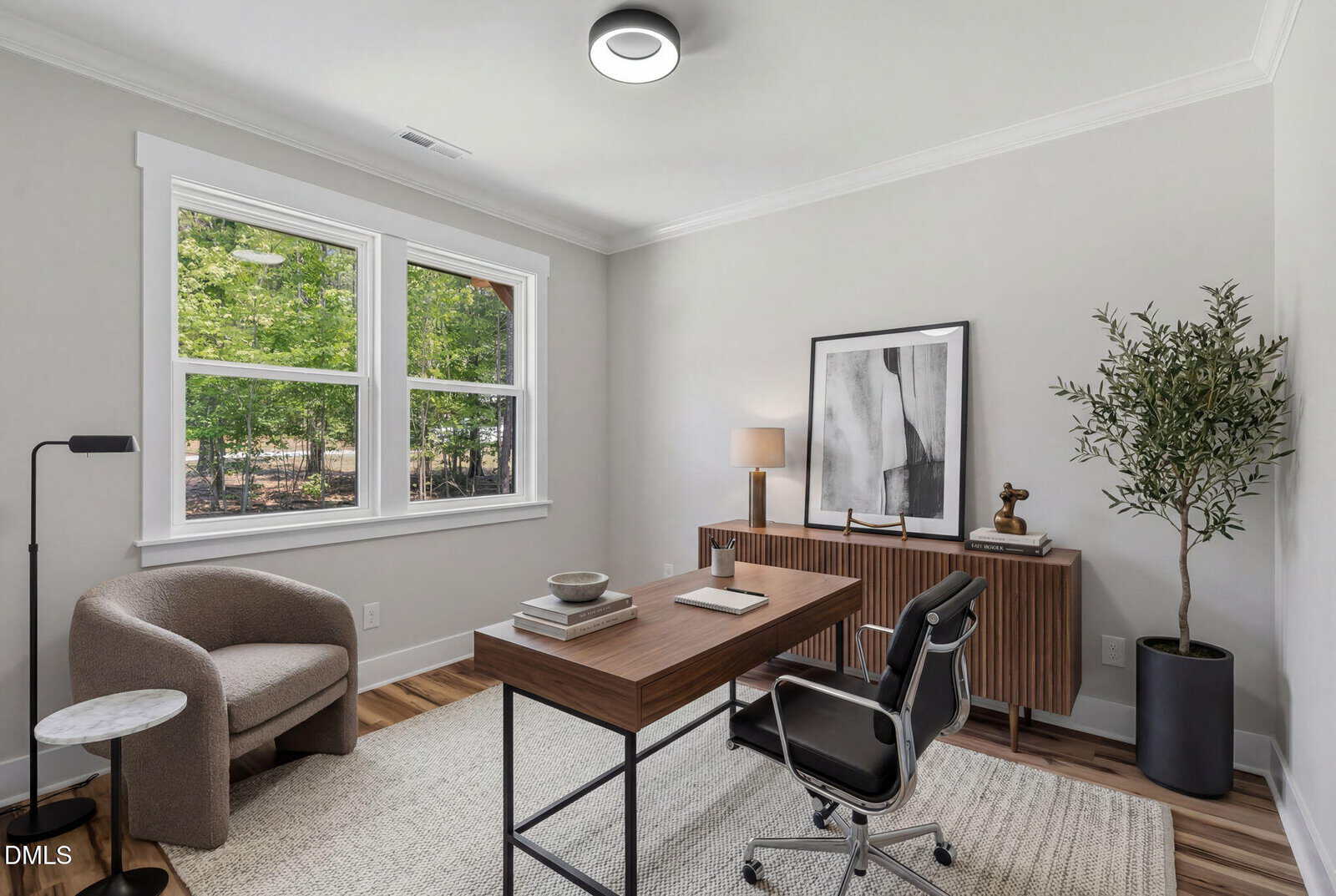 Modern home office with a wooden desk, black chair, beige armchair, side table, credenza, framed abstract art, potted tree, lamp, and a large window showing green trees outside. Light walls and wood floor with a rug.
