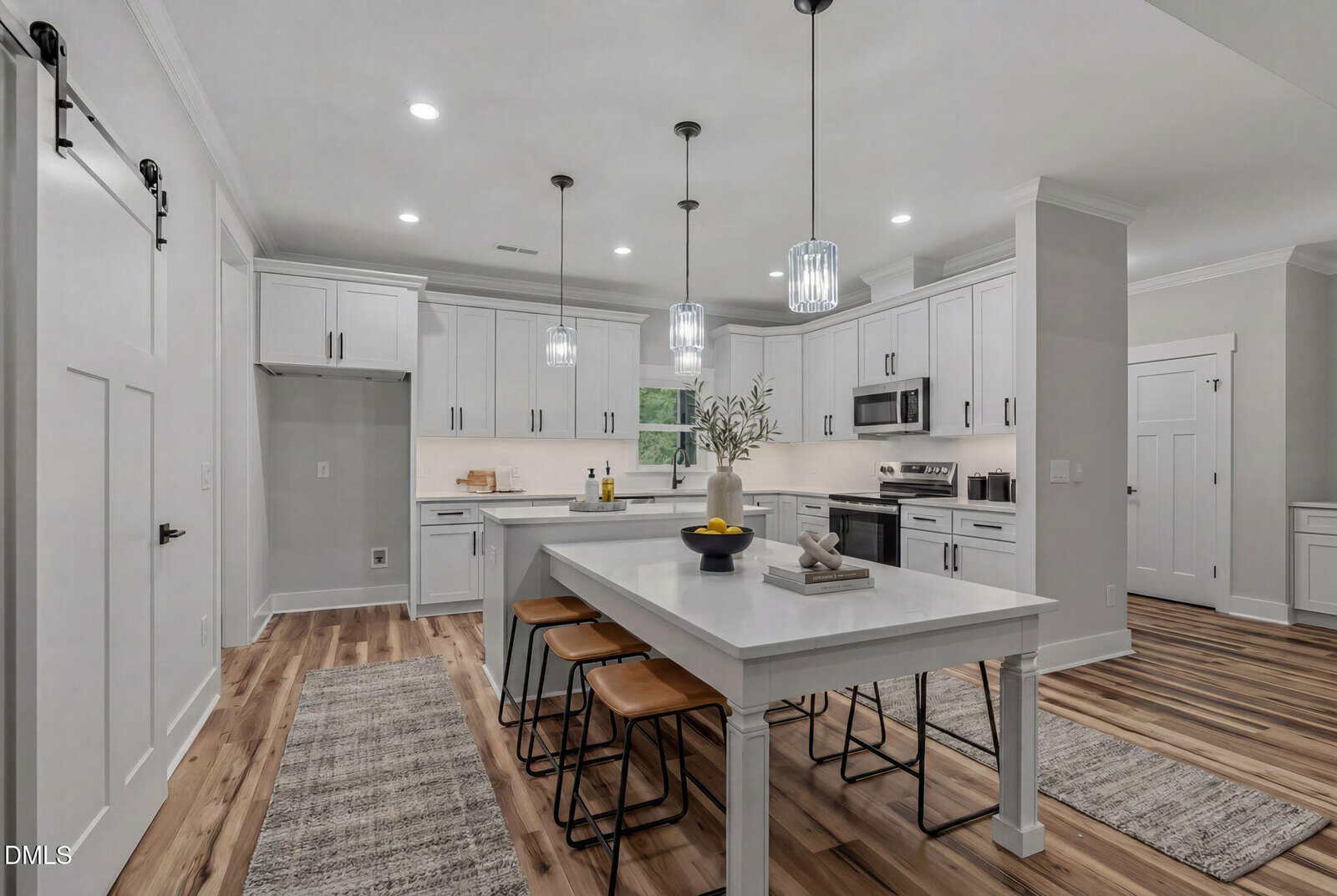 Modern kitchen with white cabinets, stainless steel appliances, large island with four wooden stools, pendant lights above, and two rugs on a wood floor. A vase and bowl with fruit are on the island.