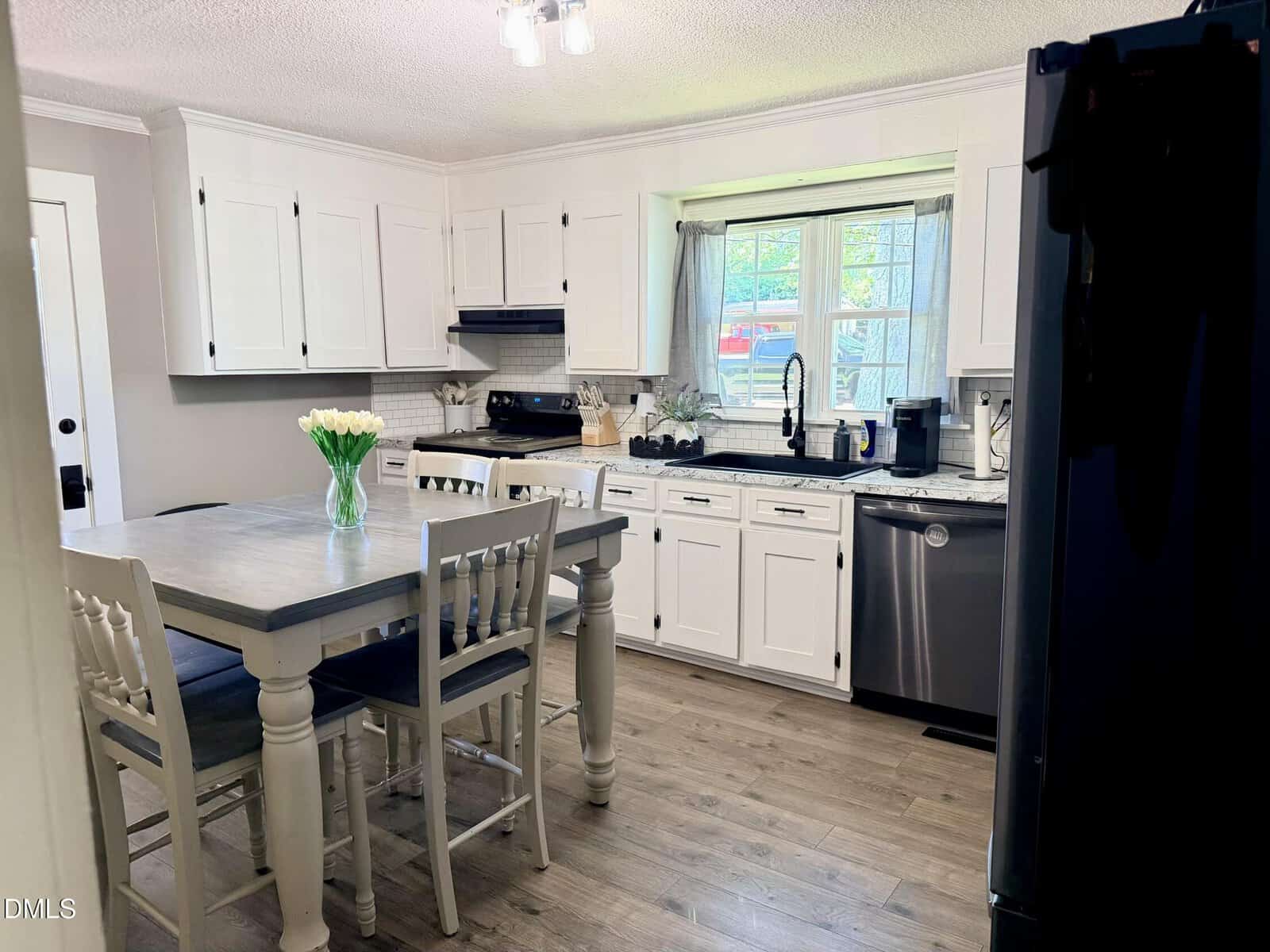 A bright kitchen with white cabinets, stainless steel appliances, a wooden table with four chairs, and a vase of white tulips. Sunlight streams in through a window above the sink.
