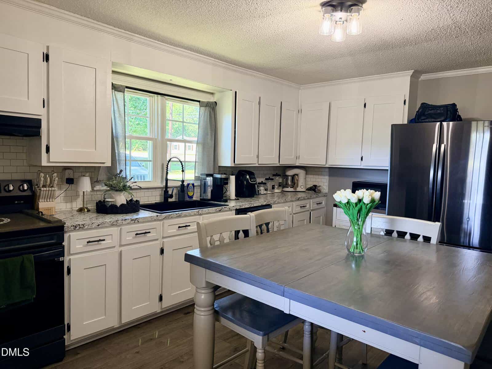 A modern kitchen with white cabinets, stainless steel appliances, a gray island table, and a vase of white tulips. Sunlight streams through the window above the sink.