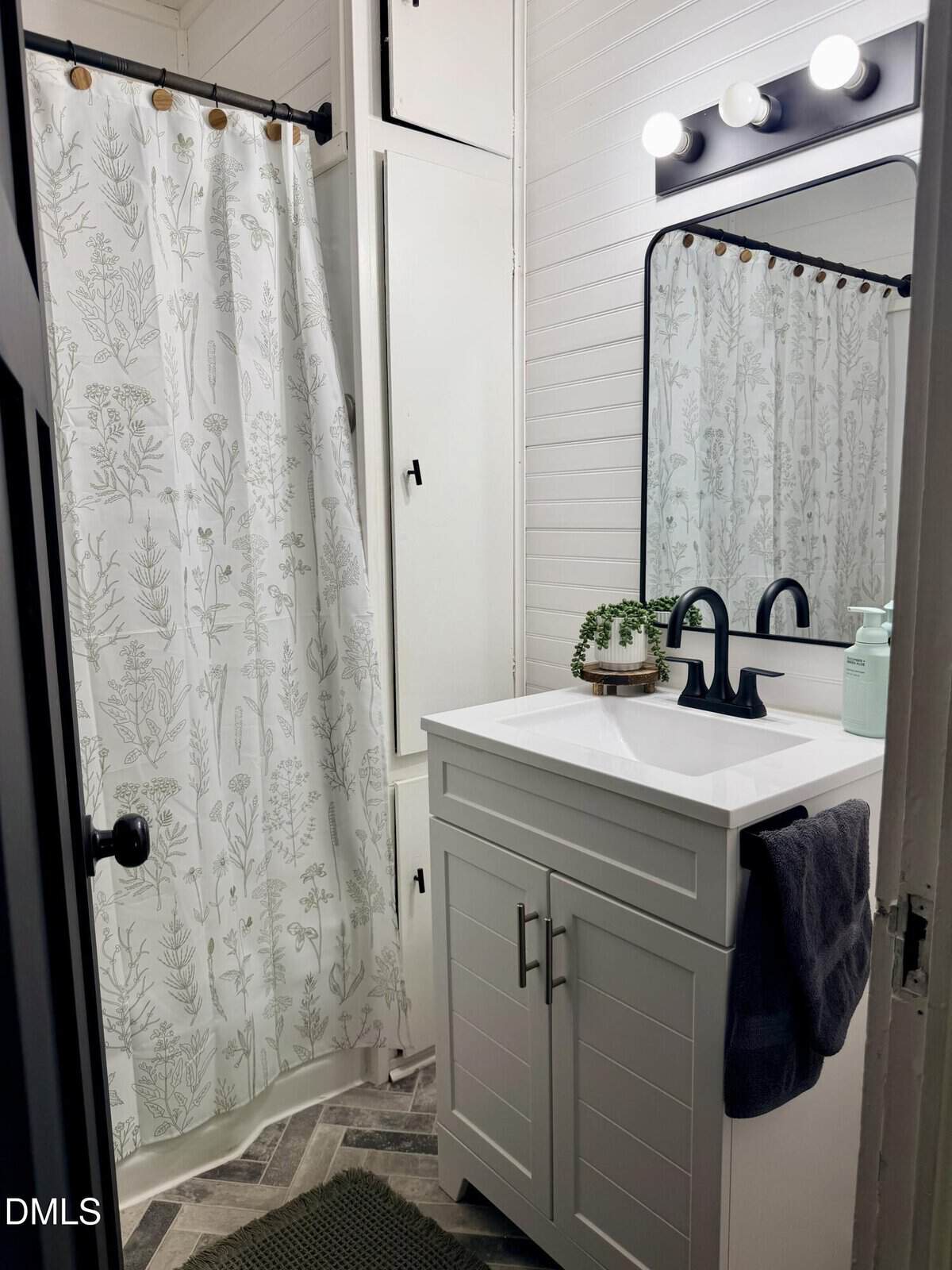 Small bathroom with a white vanity, black faucet, large mirror, potted plant, gray towel, and a shower with a botanical-patterned curtain. White walls and modern lighting complete the look.