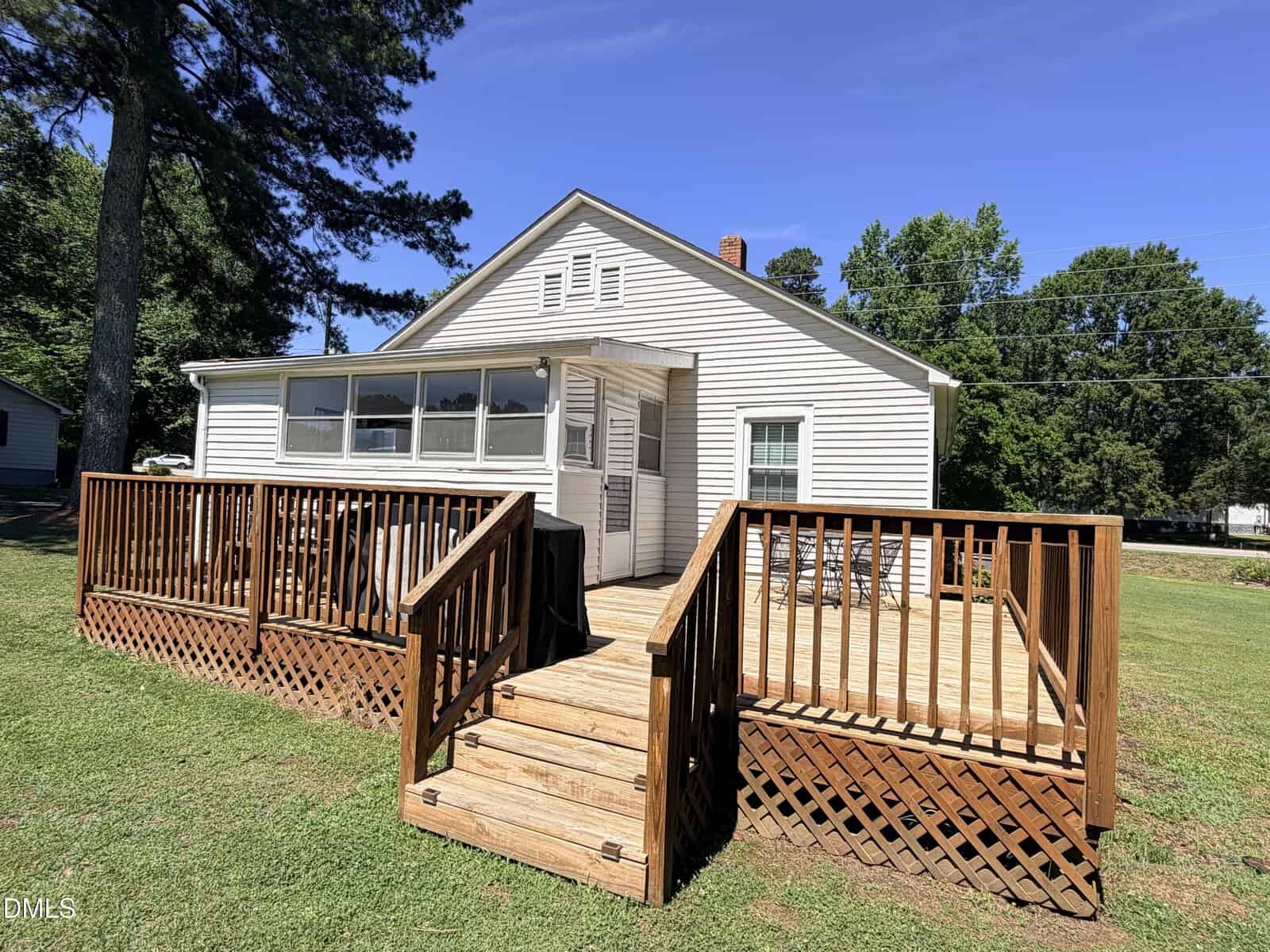 A small white house with a gabled roof and a large wooden ramp and deck leading to a sunroom entrance, surrounded by grass and trees under a clear blue sky.