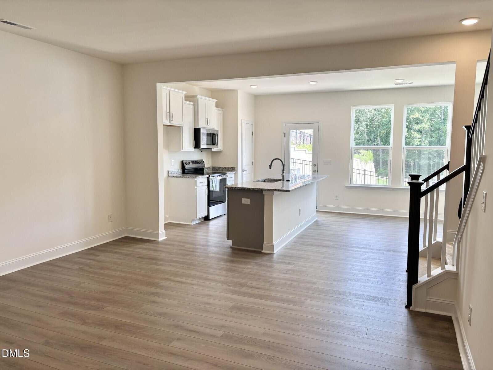 Modern open-concept living space with light wood floors, white walls, a kitchen with white cabinets and stainless steel appliances, large windows, and a staircase with a black railing.