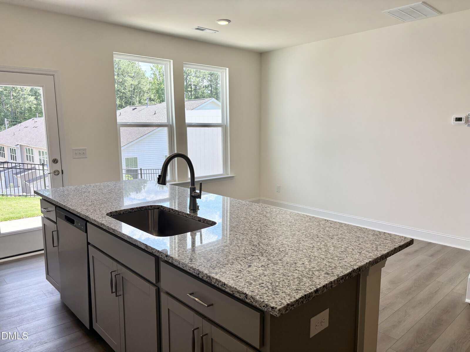 A modern kitchen with a granite island countertop, sink, stainless steel faucet, gray cabinets, wood flooring, and large windows that let in natural light. A back door opens to a grassy yard.