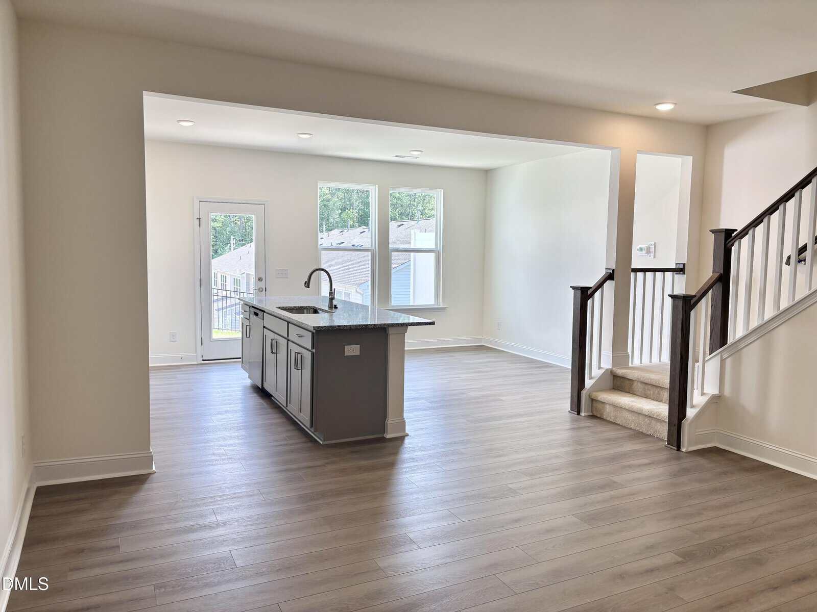 Open-concept living area with light wood flooring, a kitchen island with a sink, large windows, a door to the outside, and a staircase leading to an upper floor. Walls are white and space is well-lit by natural light.