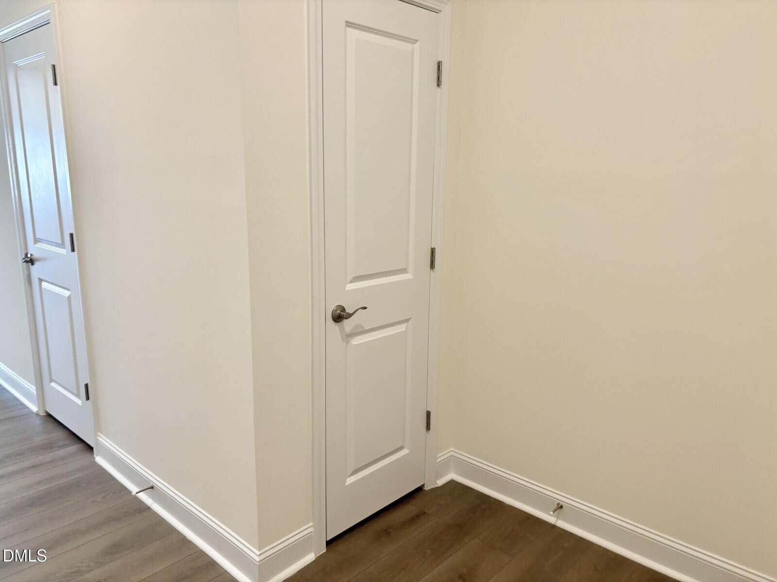 A corner of a room with beige walls, white trim, and two closed white doors. The floor is wood, and there is a white baseboard along the walls. One door is near the corner, the other is in the hallway.