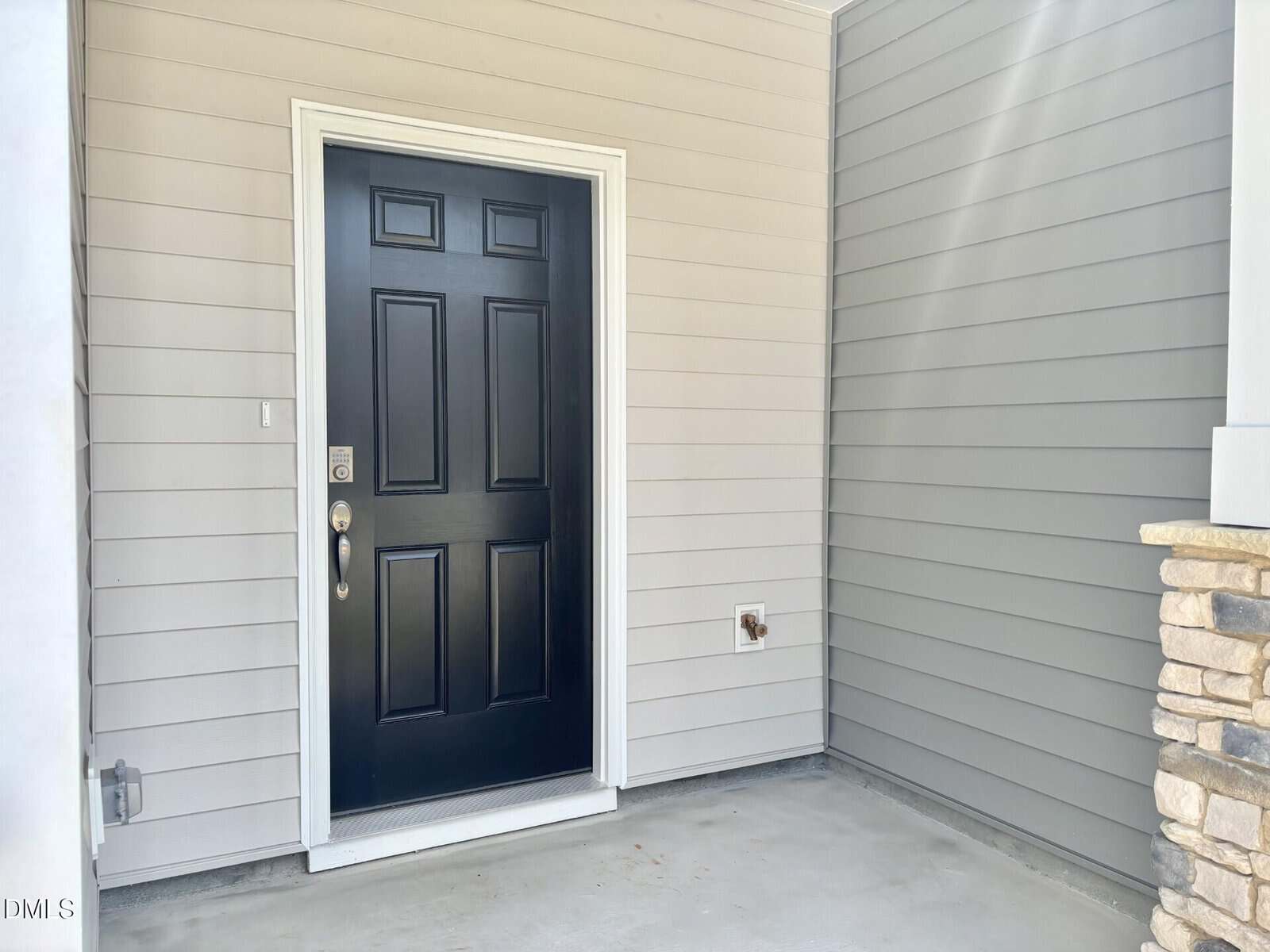 A black front door with a silver handle and keypad is set in light gray horizontal siding. A covered concrete entryway has a white trim and a partial stone pillar on the right.
