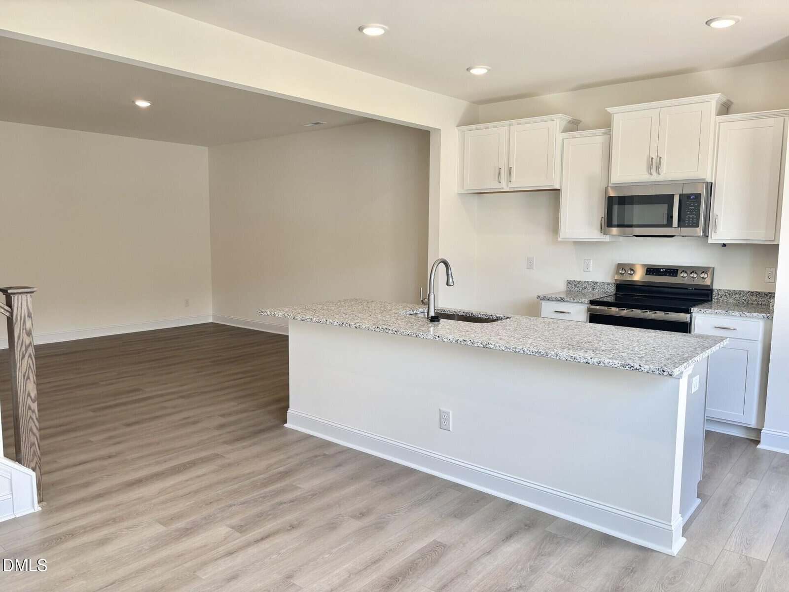 Modern kitchen with white cabinets, stainless steel appliances, granite countertops, and a central island with a sink. Open layout connects to a spacious living area with light wood flooring and neutral walls.