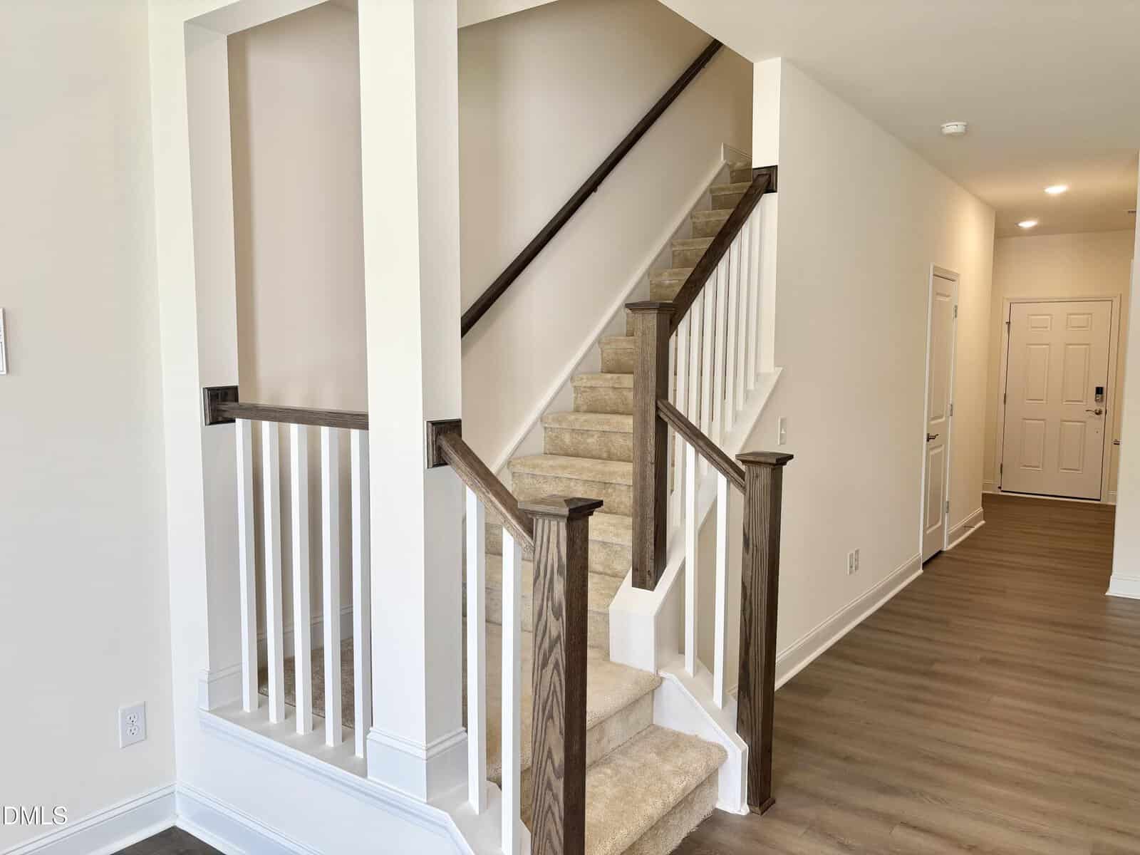A carpeted staircase with dark wood handrails and white spindles in a bright hallway with light-colored walls and wood floors. A white door is visible at the end of the hallway.