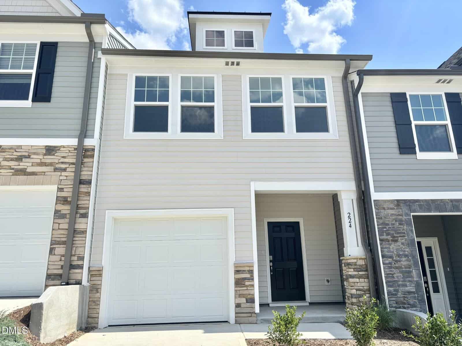 This two-story townhouse at 224 Alberta Drive in Franklinton features beige siding, a black front door, and a single-car garage. The home is enhanced by multiple large windows and elegant stone accents on the lower left and right sides. Above, the sky is partly cloudy, providing a serene backdrop to this inviting residence.