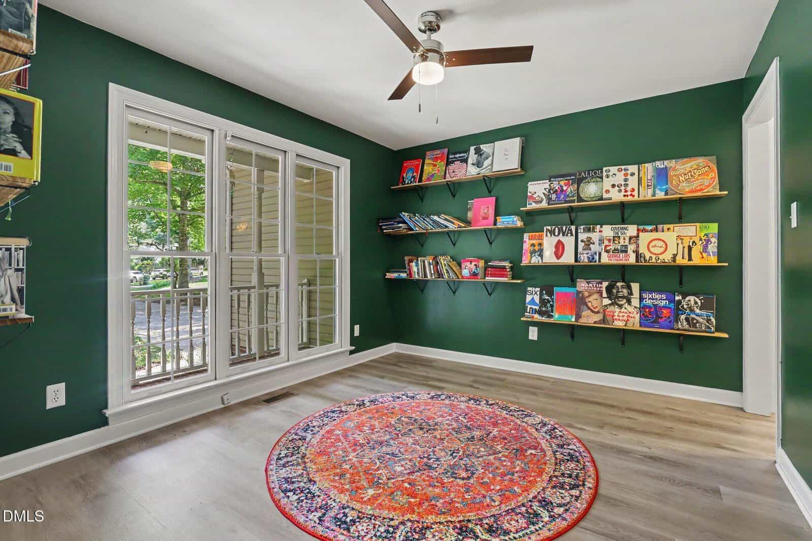 A bright room with dark green walls, a ceiling fan, large windows, floating shelves holding books and records, and a colorful round rug on light wood flooring.
