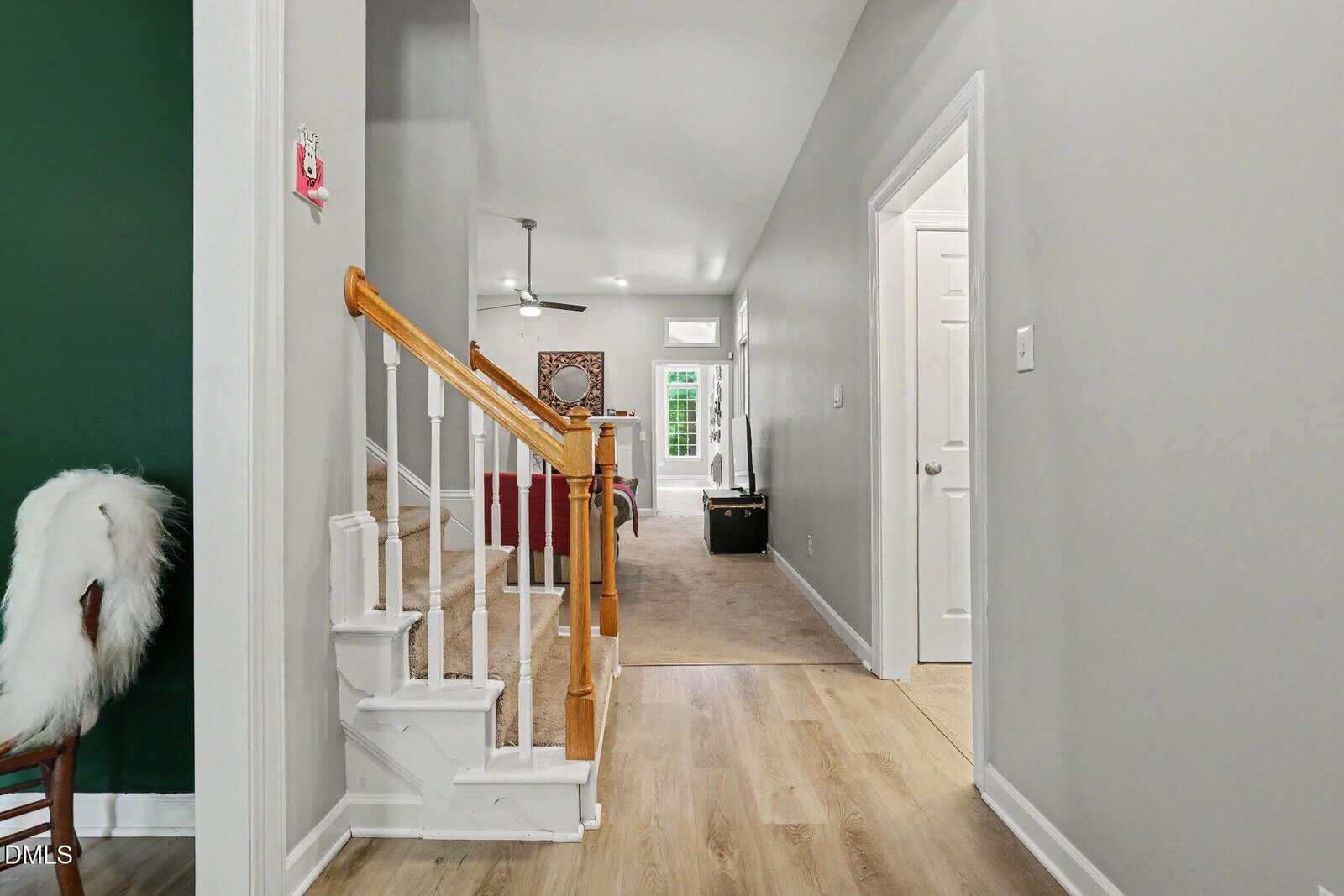 A hallway with light wood flooring leads to a carpeted living area. A white staircase with wooden handrail is on the left, and a white fur throw drapes over a chair against a green wall. Doors line the right side of the hallway.