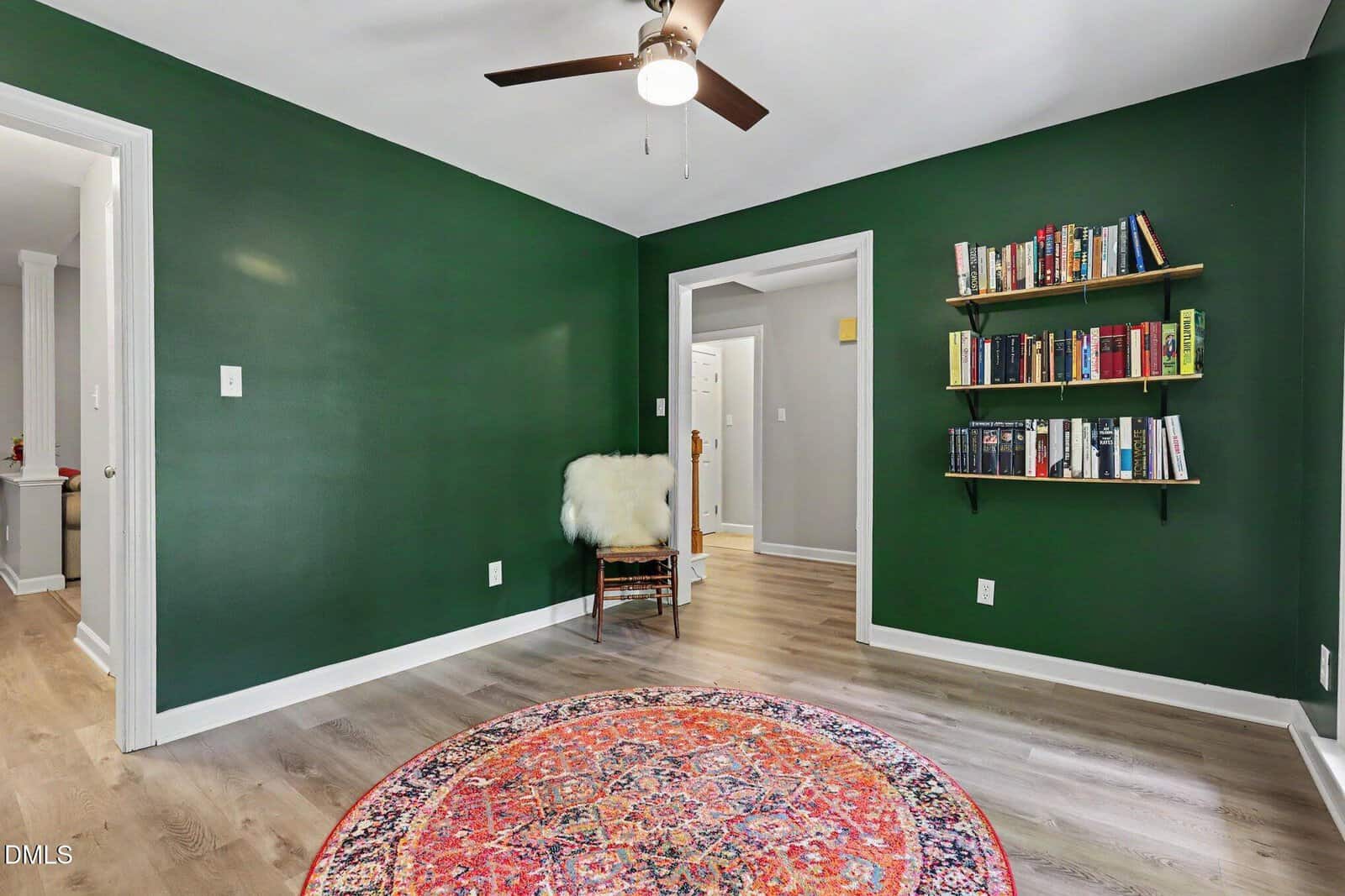A room with dark green walls, light wood flooring, a ceiling fan, a circular patterned rug, a chair with a white fur throw, and two black floating bookshelves filled with books. Doorways lead to adjacent rooms.