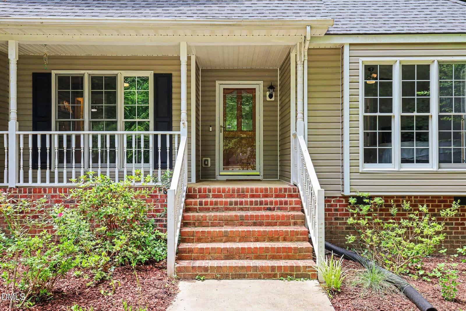 Front porch of a house with brick steps, white railings, beige siding, black shutters, and large windows. There are green shrubs and mulch in the garden beds along the walkway.