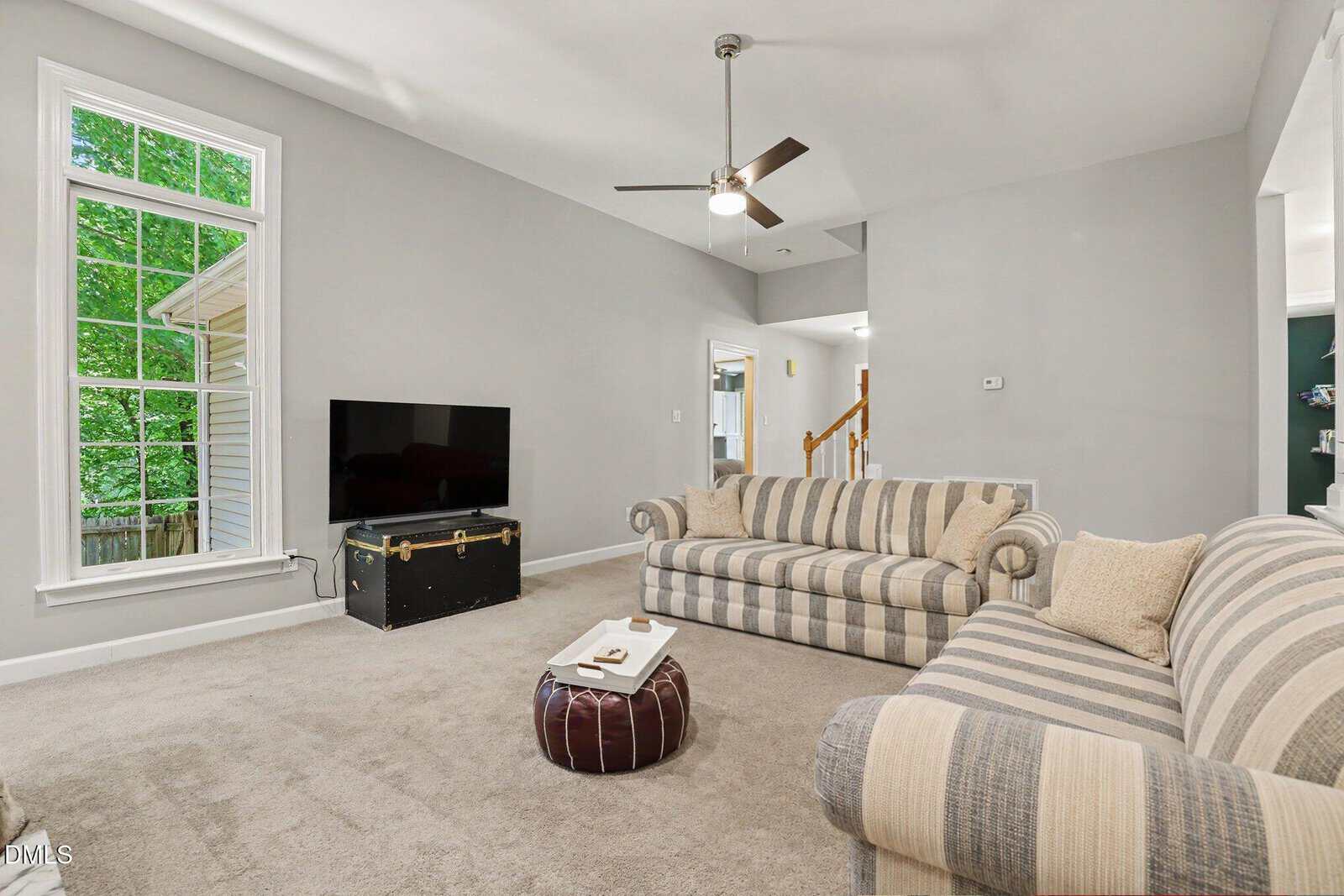 A bright living room with two striped sofas, a TV on a black chest, a brown ottoman, beige carpet, gray walls, a large window with greenery outside, and a ceiling fan.