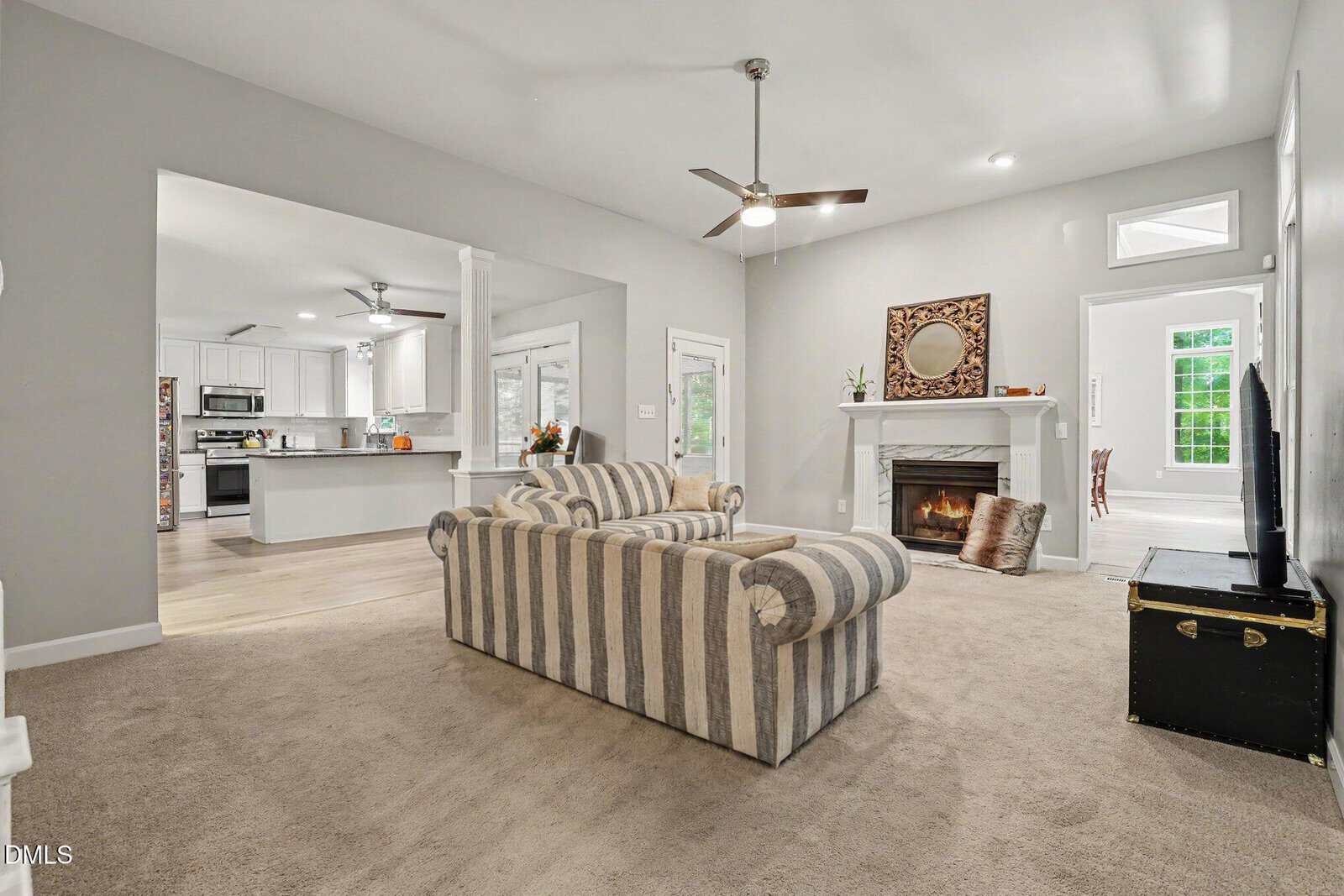 A cozy living room with a striped sofa, a fireplace with a mirror above it, a TV on a trunk, and an open view into a modern white kitchen and a bright dining area.