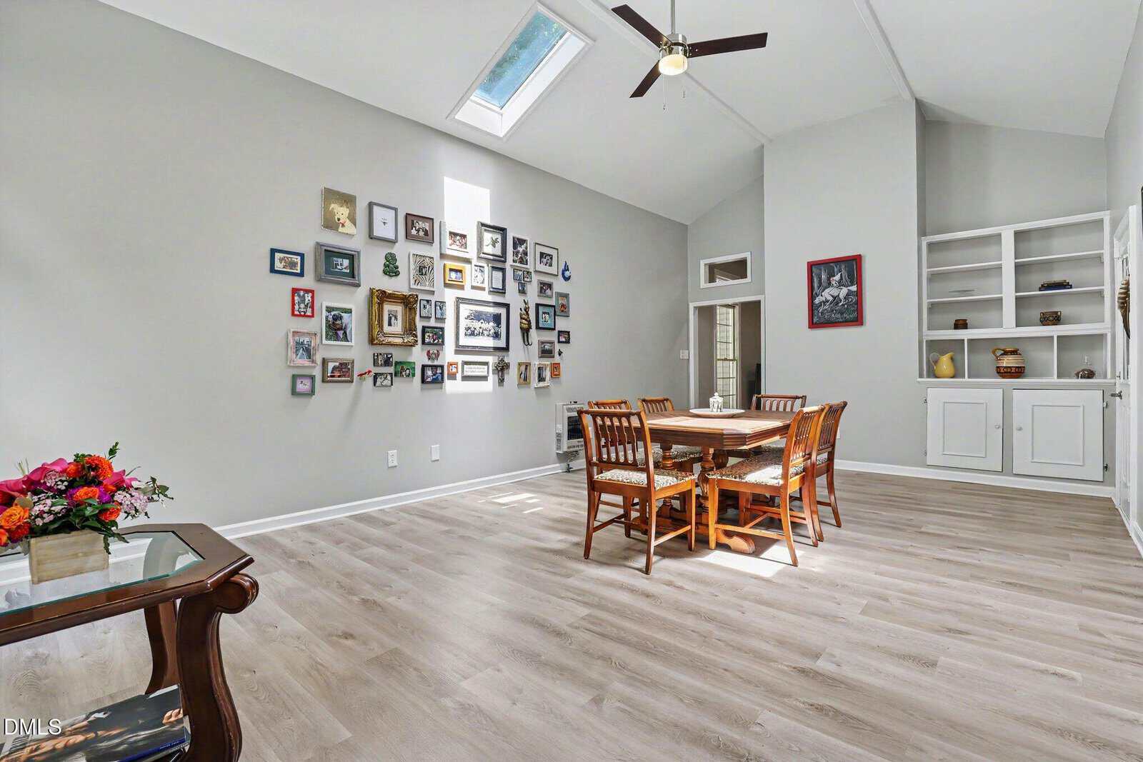 Bright dining room with light wood floors, a large skylight, a ceiling fan, a wooden table with chairs, a wall of various framed photos, white built-in shelves, and a side table with a colorful flower arrangement.