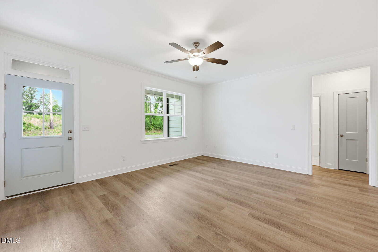 Bright, empty room with light wood flooring, white walls, a ceiling fan with lights, gray door, large window, and an open doorway leading to another room.