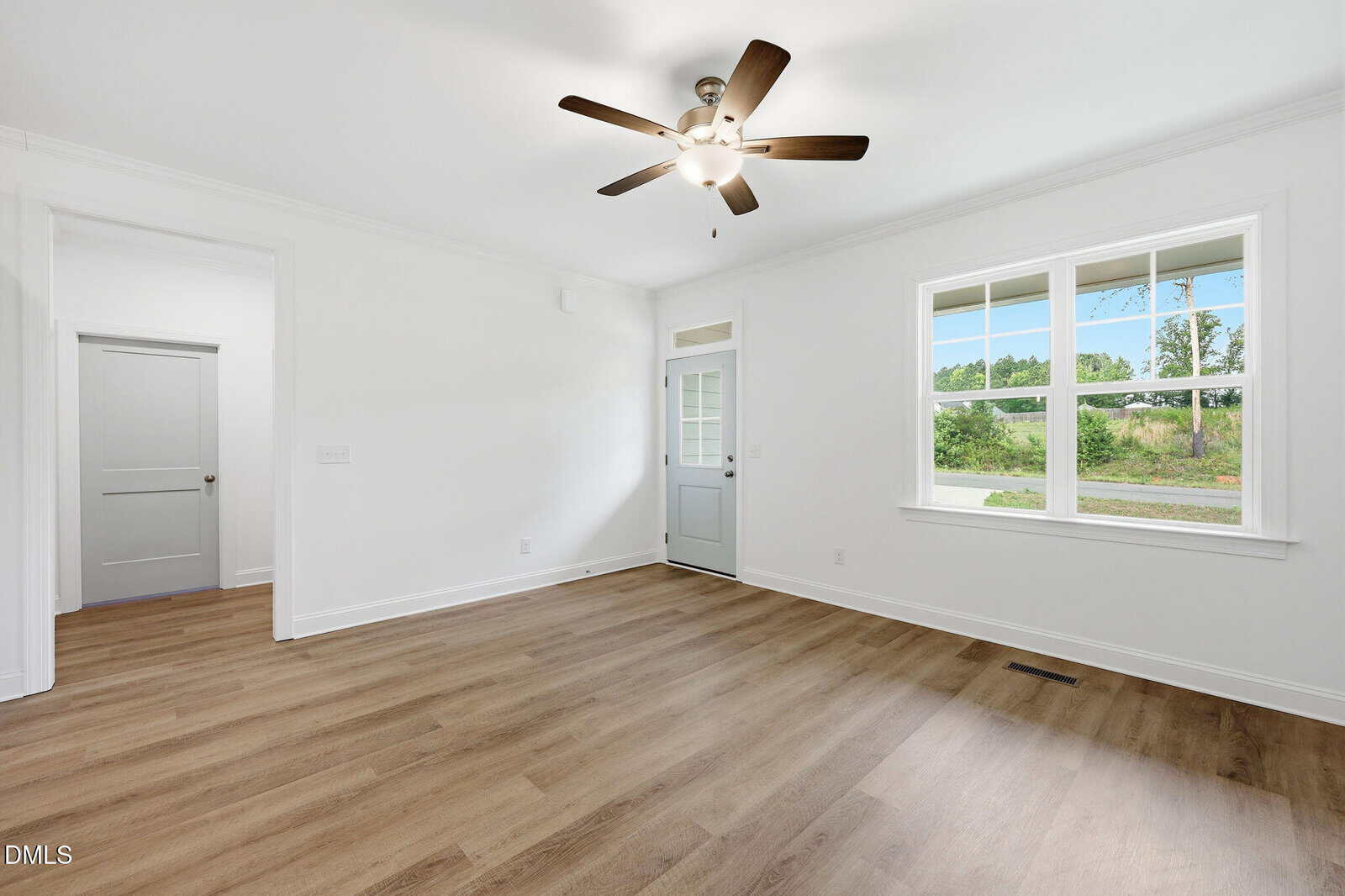 Bright empty room with wood floors, white walls, a ceiling fan, a large window showing greenery outside, and a light gray door with glass panes.