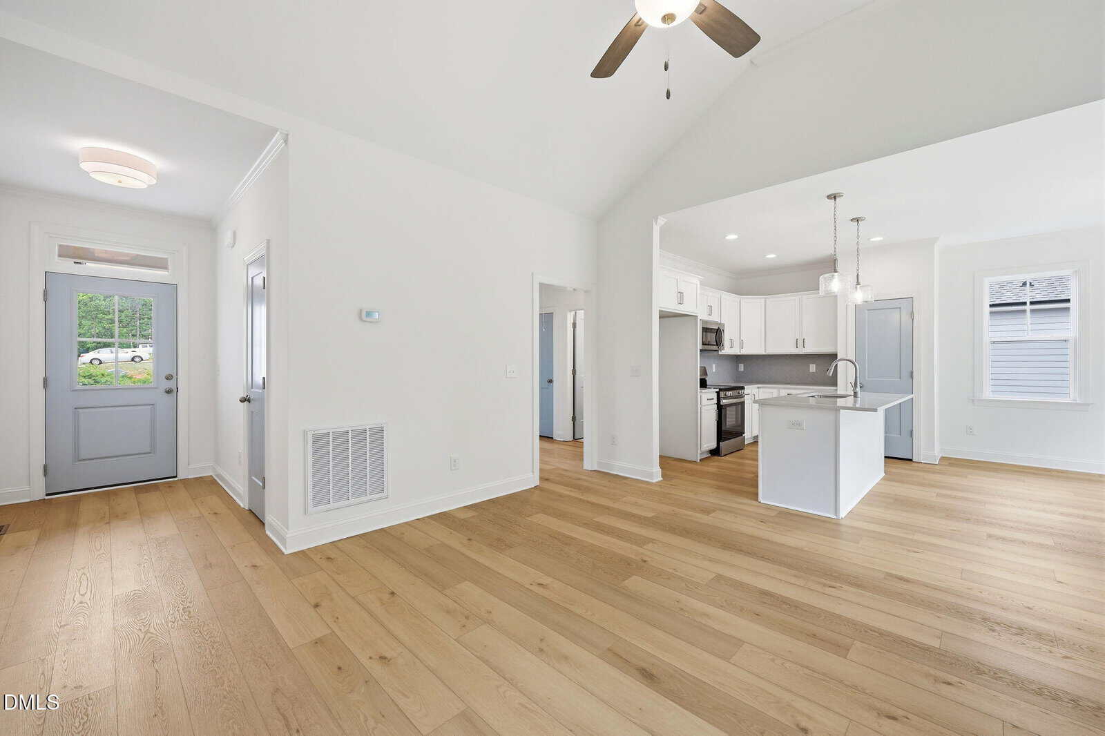 Bright, modern open-concept living area with light wood floors, white walls, a ceiling fan, and a view of a kitchen with white cabinetry, pendant lights, and an island. Entryway and doors are visible.
