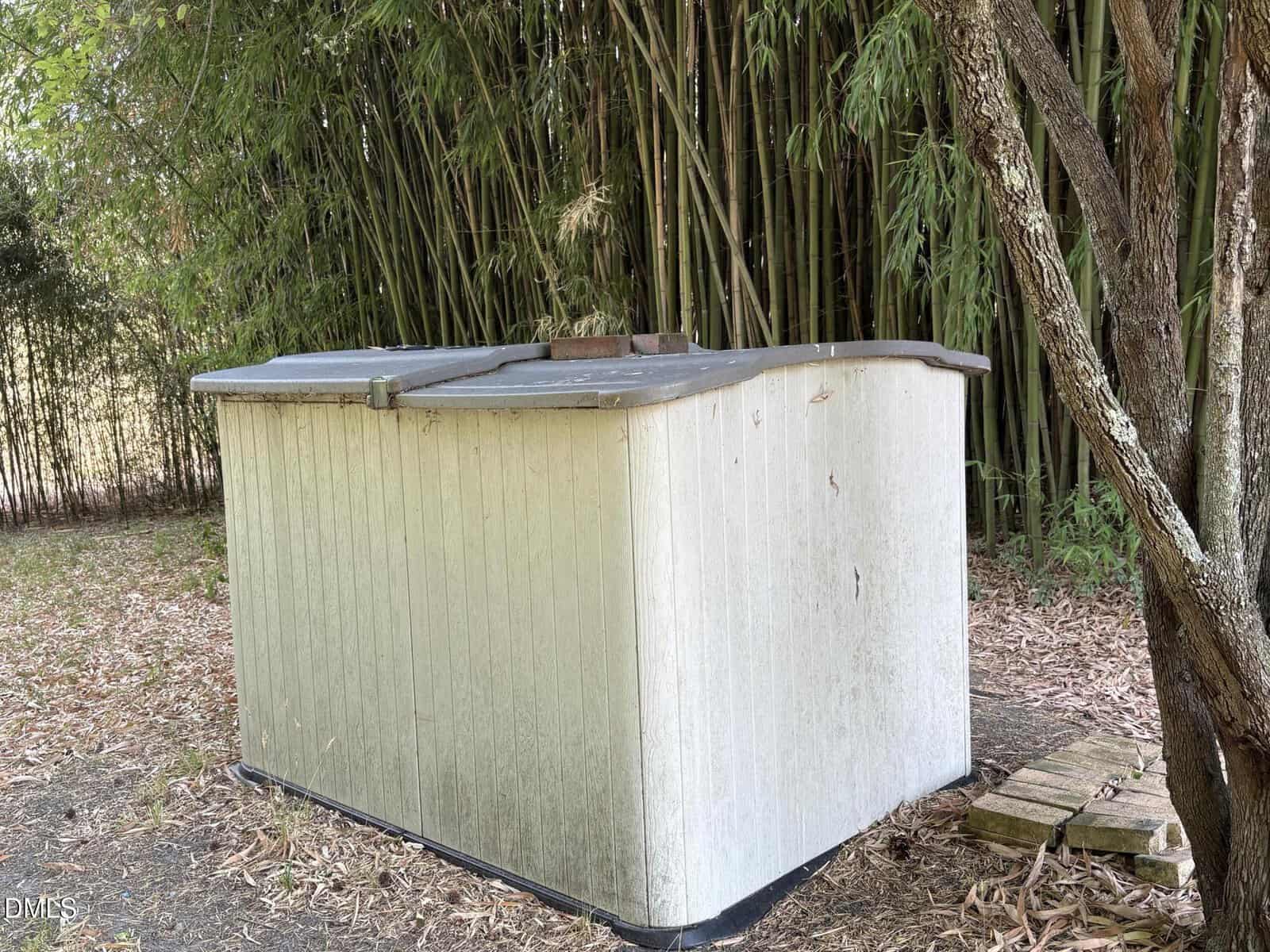 A white outdoor plastic storage shed with a dark lid stands near tall bamboo and a tree, surrounded by fallen leaves and wooden planks on the ground.