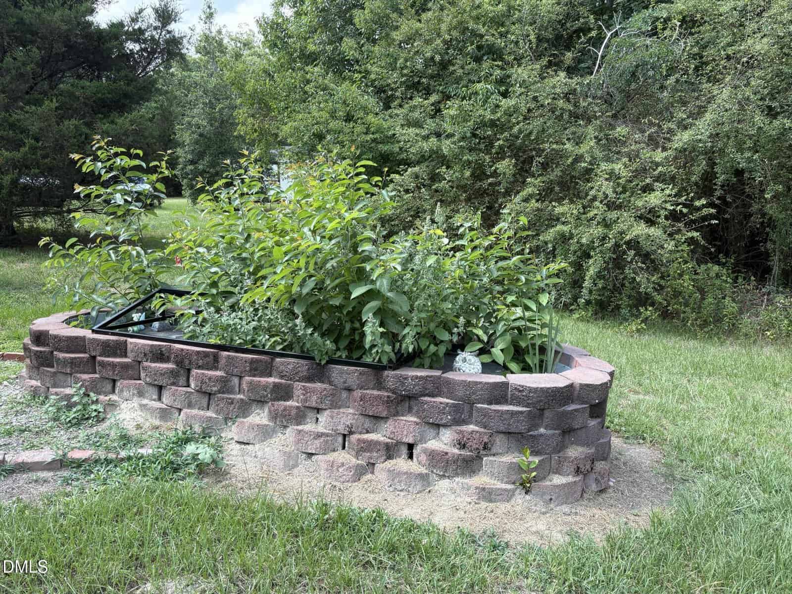A raised garden bed made of stacked stone bricks is filled with lush green plants. The bed sits on grass, surrounded by trees and bushes in a natural, outdoor setting.