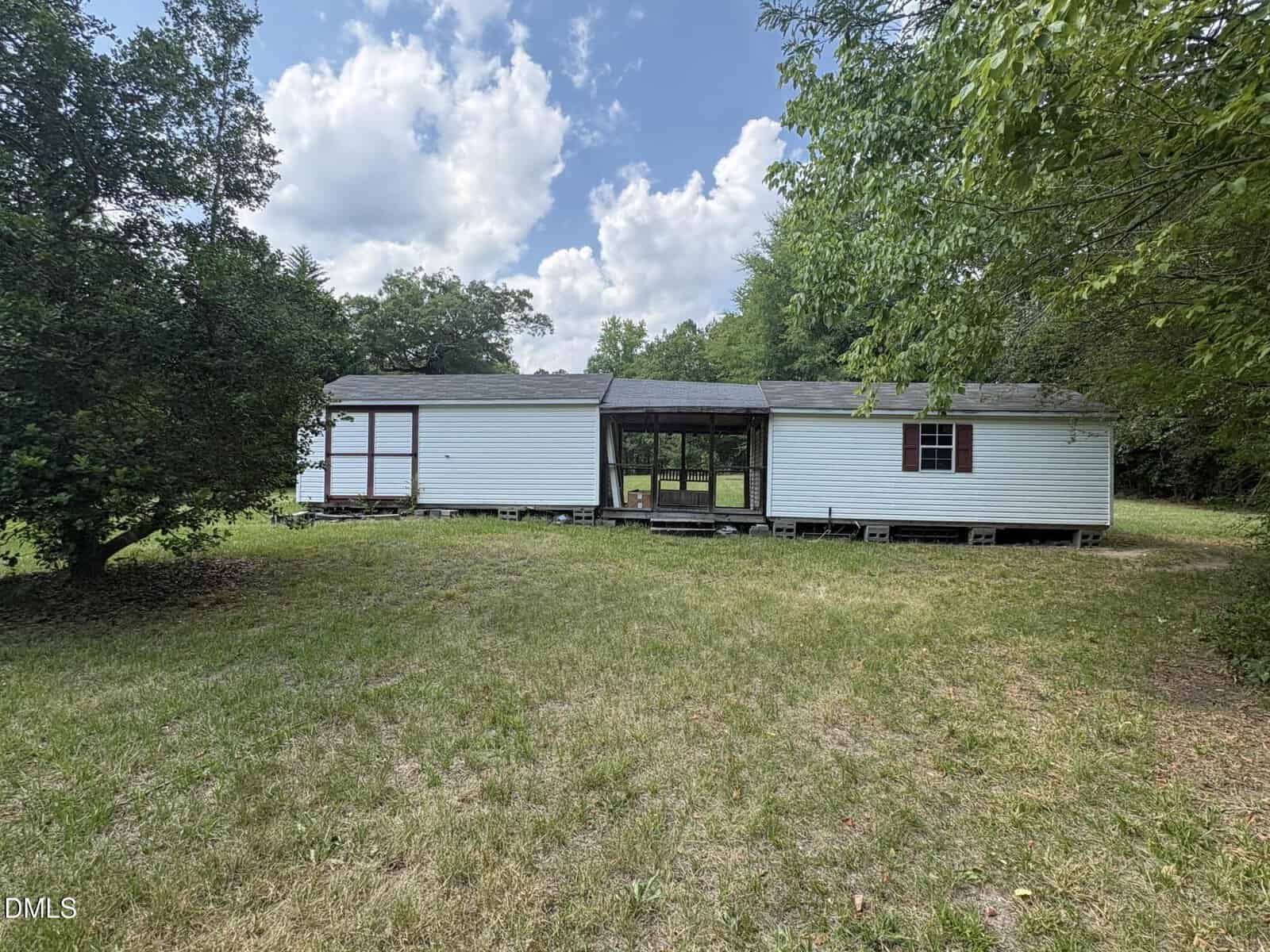A white mobile home with a screened porch sits on a grassy lot surrounded by trees under a partly cloudy sky. The home appears elevated off the ground with some visible gaps below.