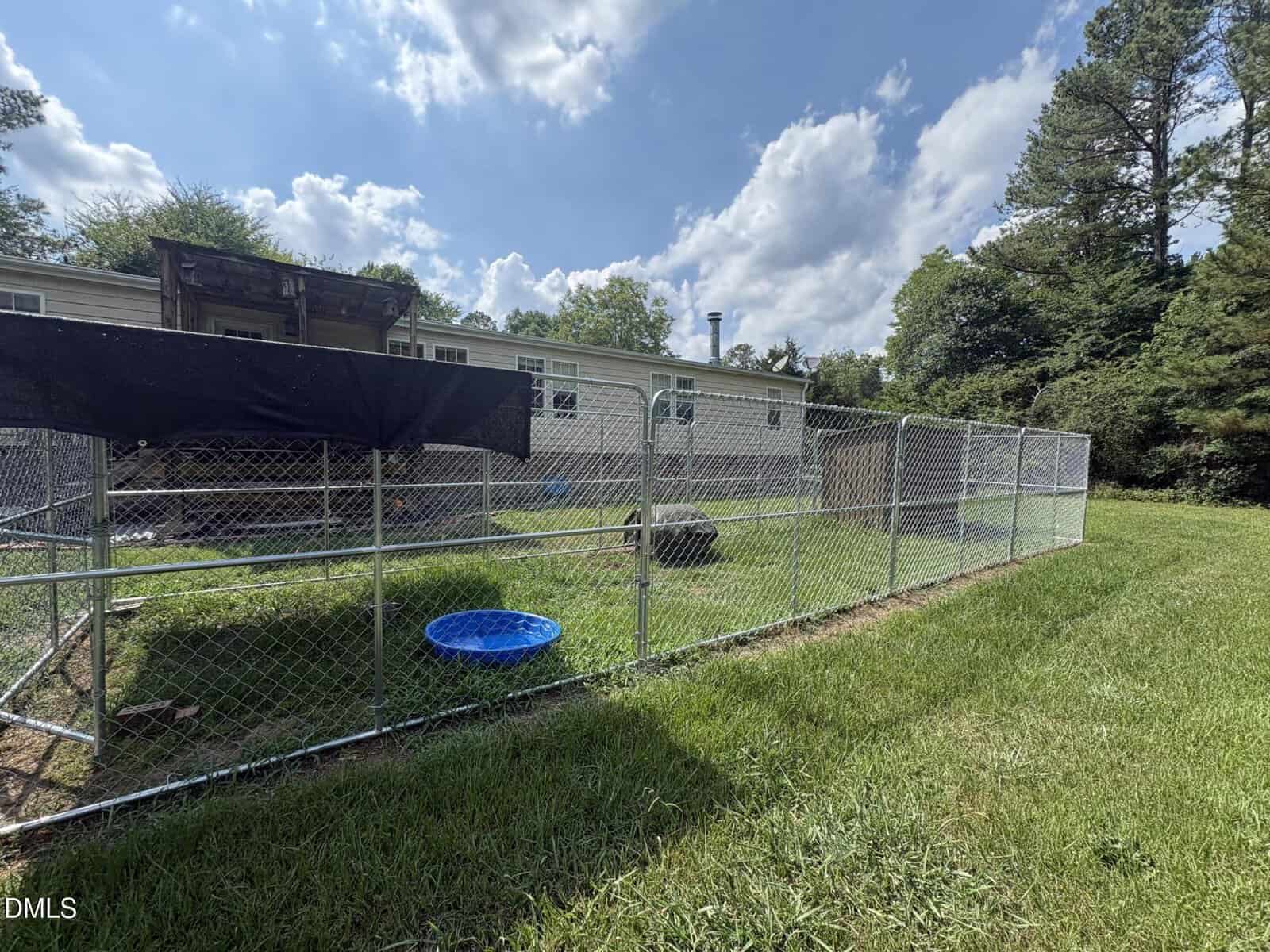 A fenced outdoor area with a small blue plastic pool, tire, and covered section beside a beige mobile home. Green grass and trees surround the yard under a partly cloudy sky.