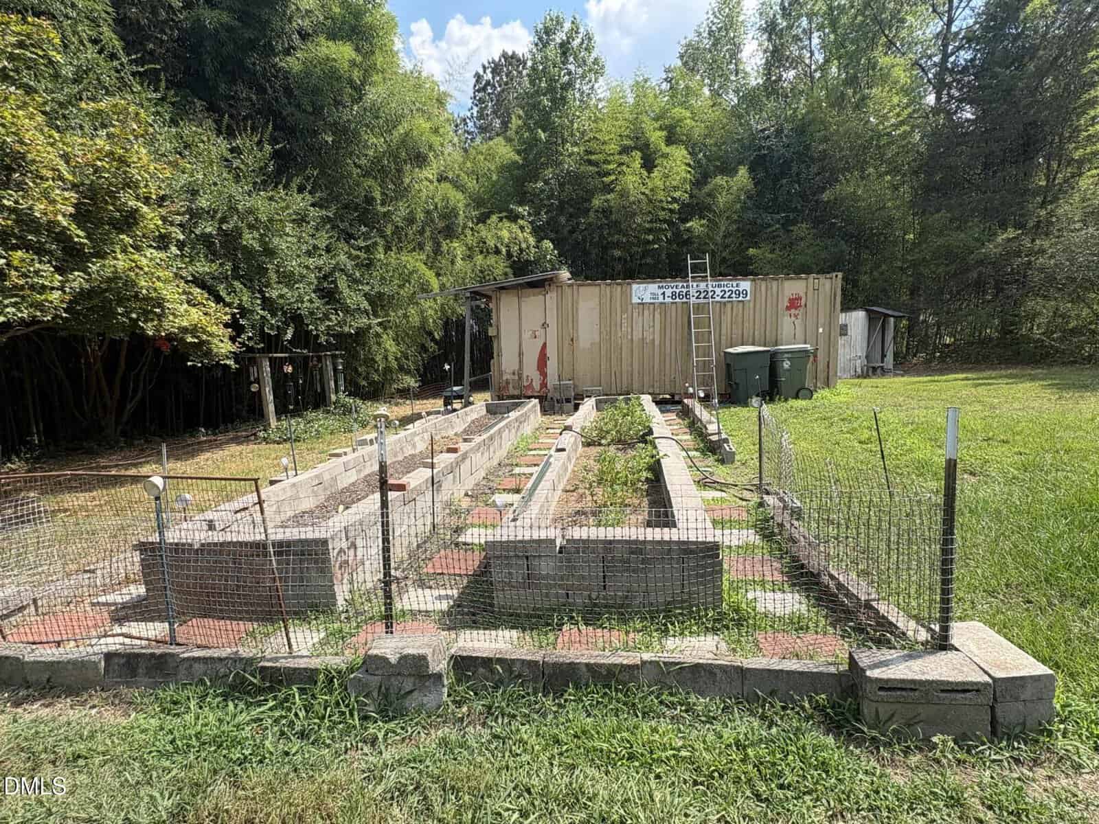 A fenced garden with raised brick beds sits on grass near a tan shipping container, surrounded by trees. A ladder leans on the container and there are trash bins nearby. The sky is partly cloudy.